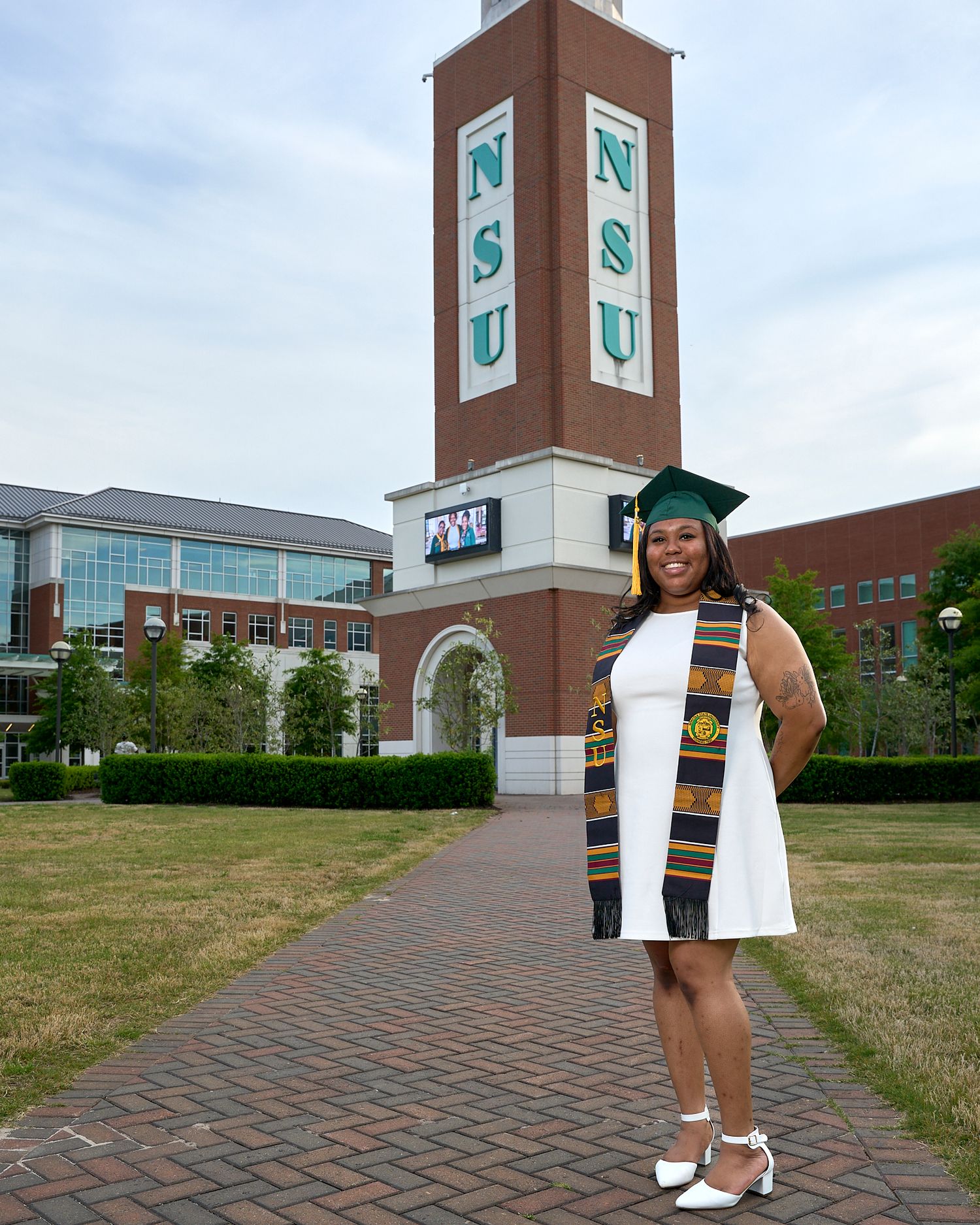 Person in white outfit poses on brick pathway near modern university building with clock tower.