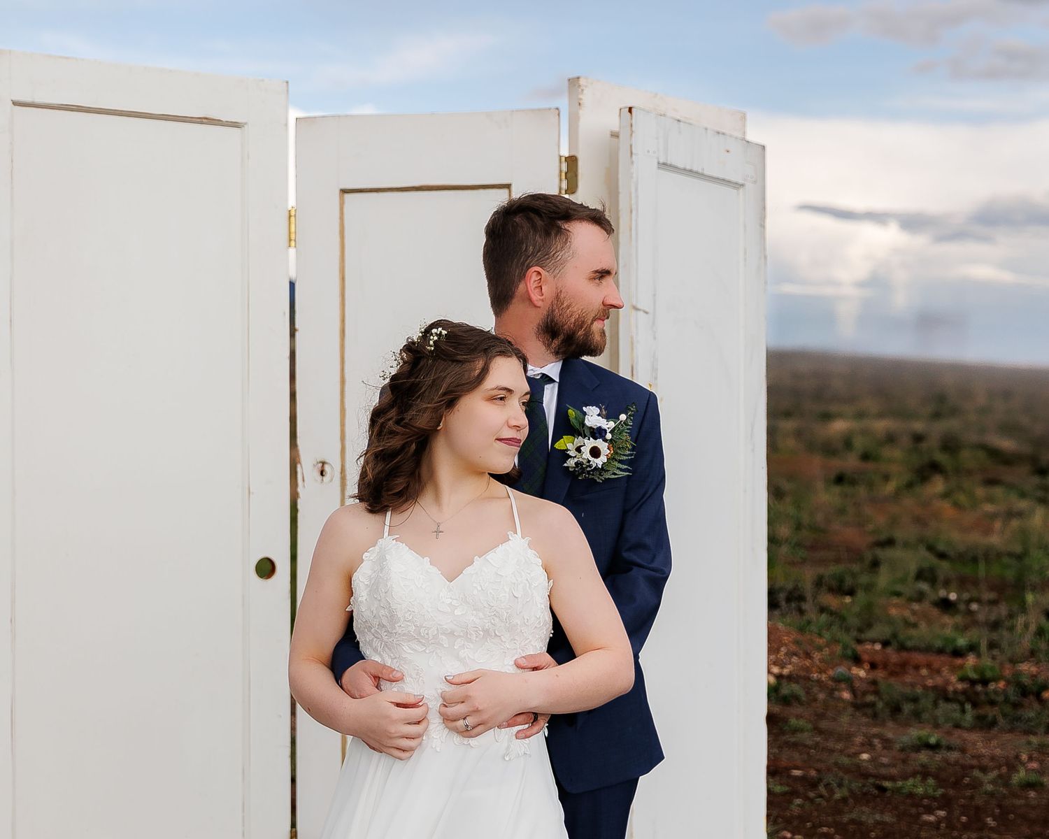 A romantic wedding couple embraces against a rustic outdoor backdrop with rolling hills and a white door.