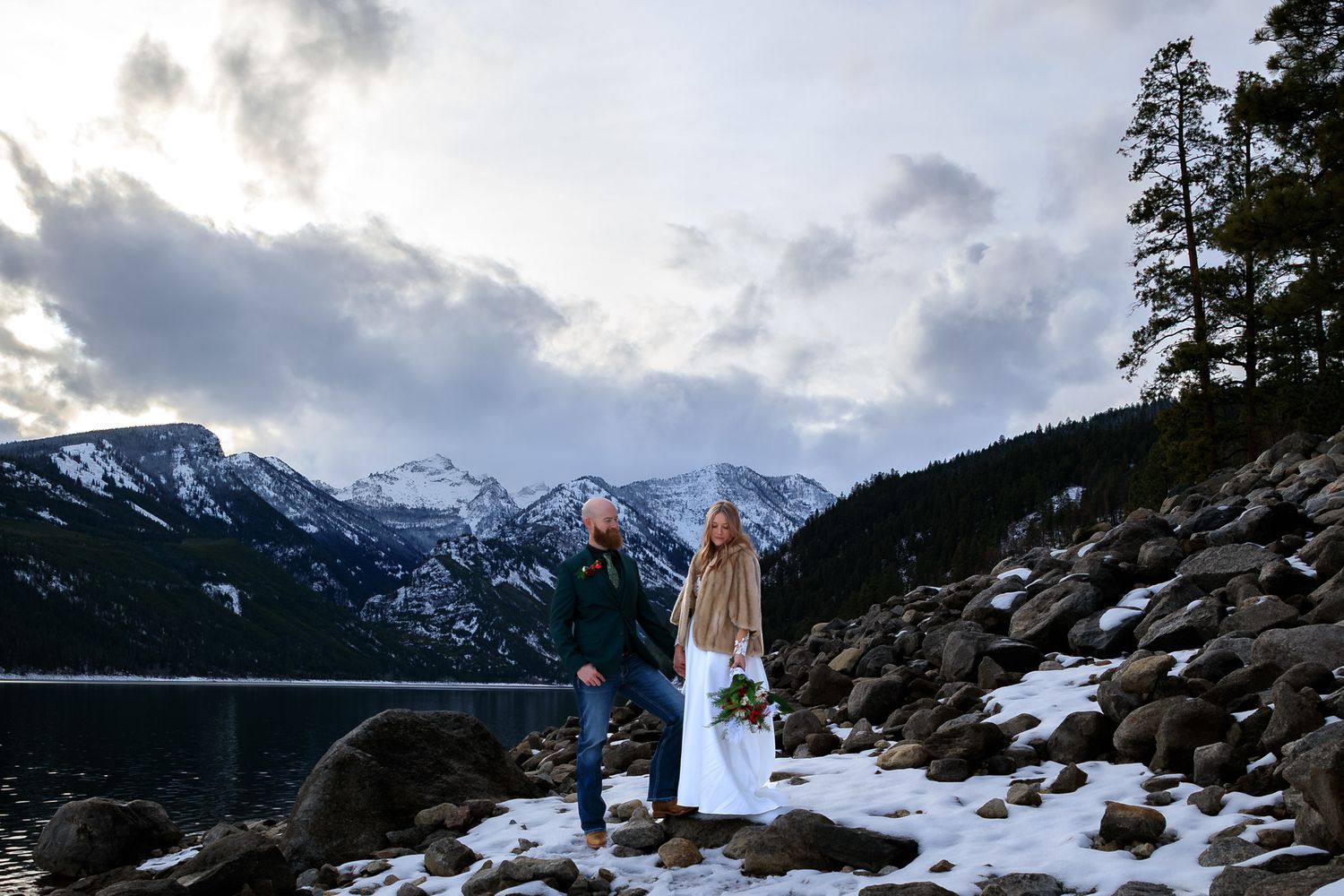 A scenic winter panorama showing snow-covered mountain peaks and evergreen trees against a cloudy sky.