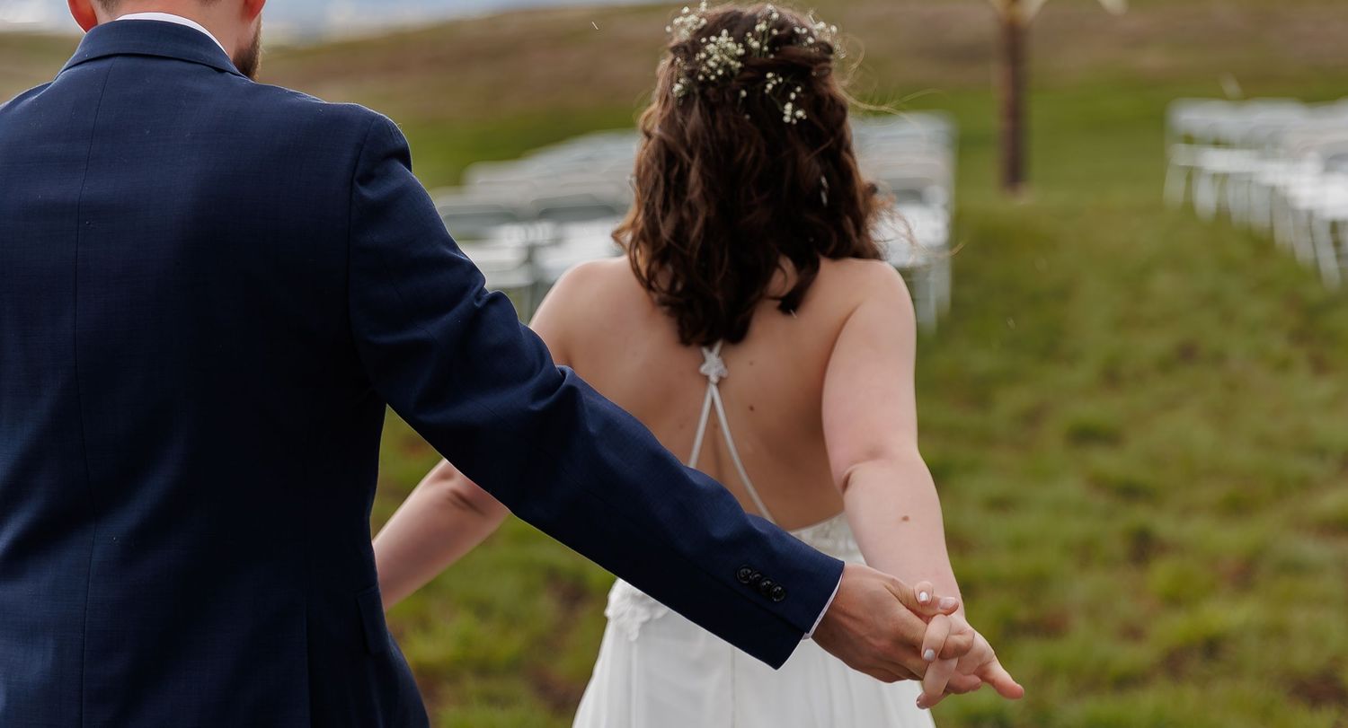 A couple in formal wedding attire holds hands while walking through a green hillside landscape.