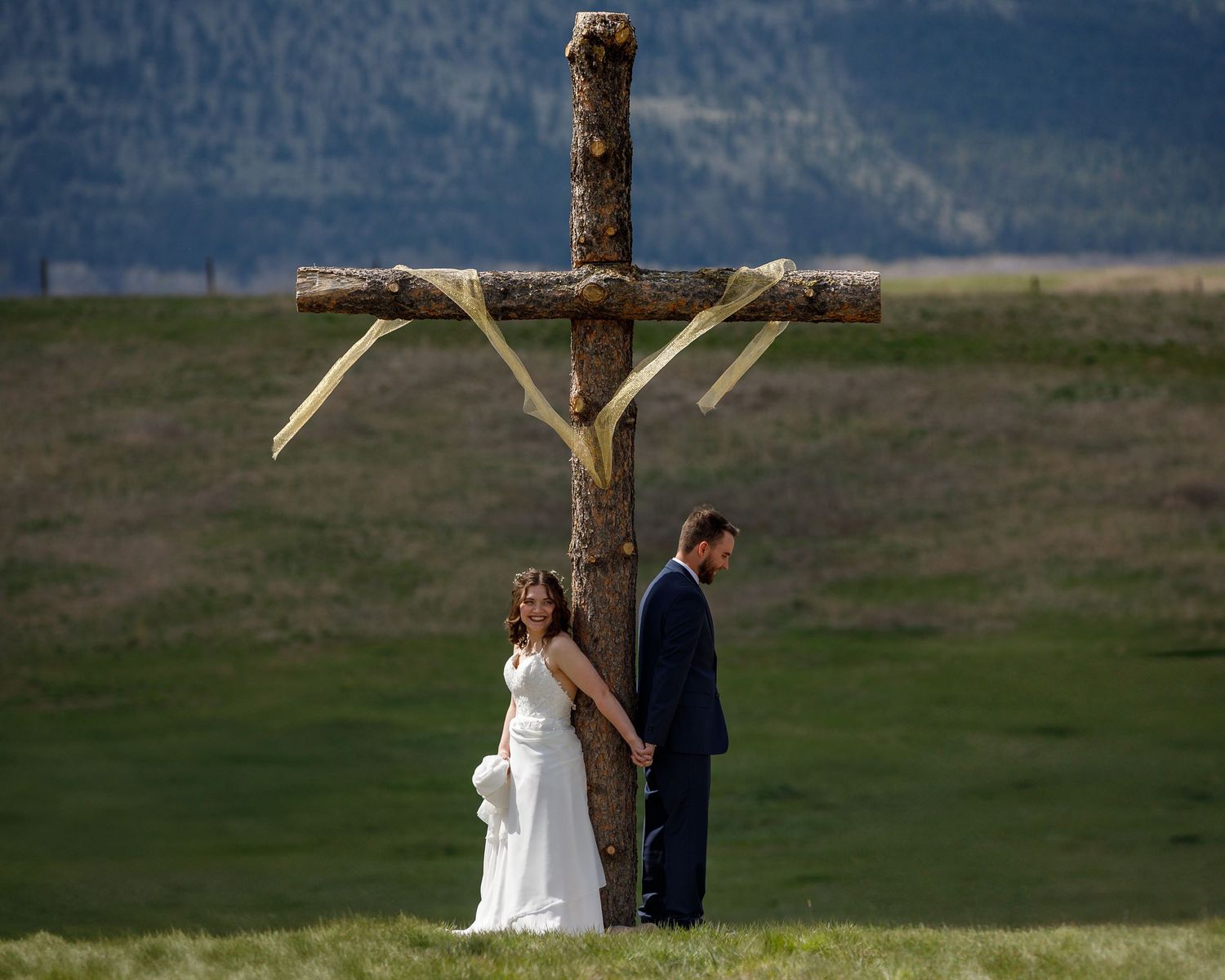 A couple in wedding attire stands together in a scenic mountain field during their outdoor ceremony.