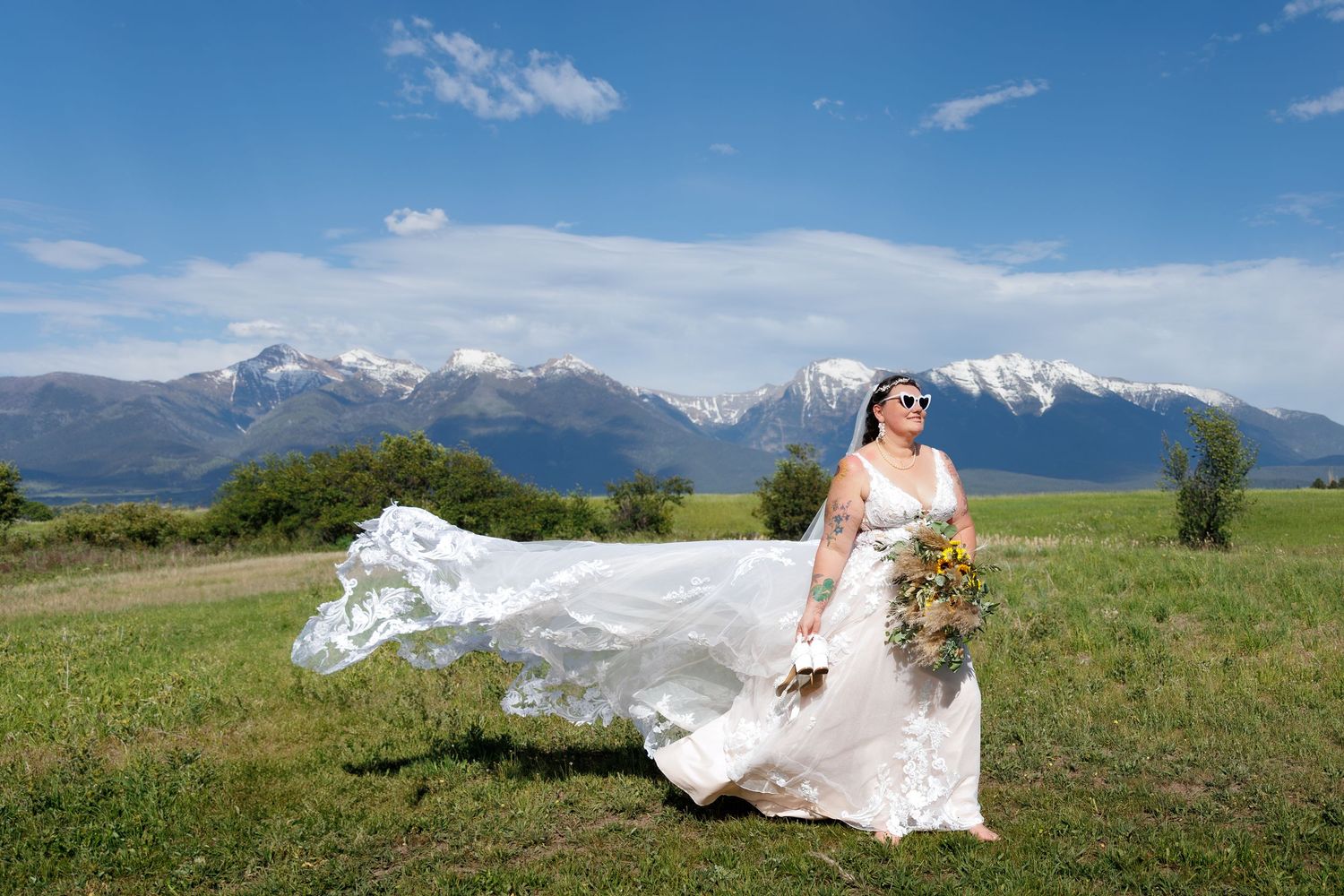 A flowing white wedding dress billows in the wind against a stunning backdrop of snow-capped mountains and green meadow at St. Ignatius