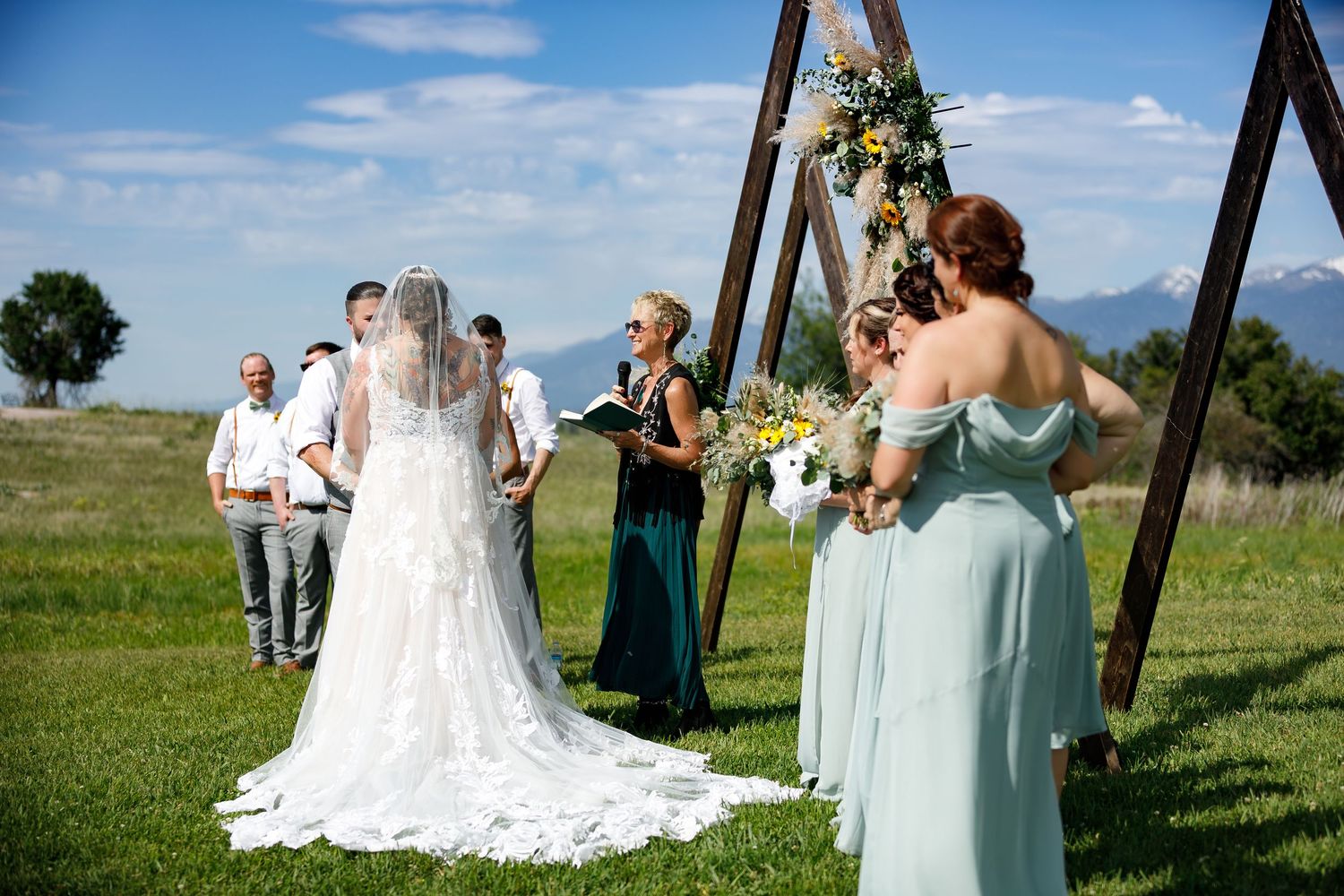Wedding party in mint green dresses poses outdoors on a grassy hill against a bright blue sky.