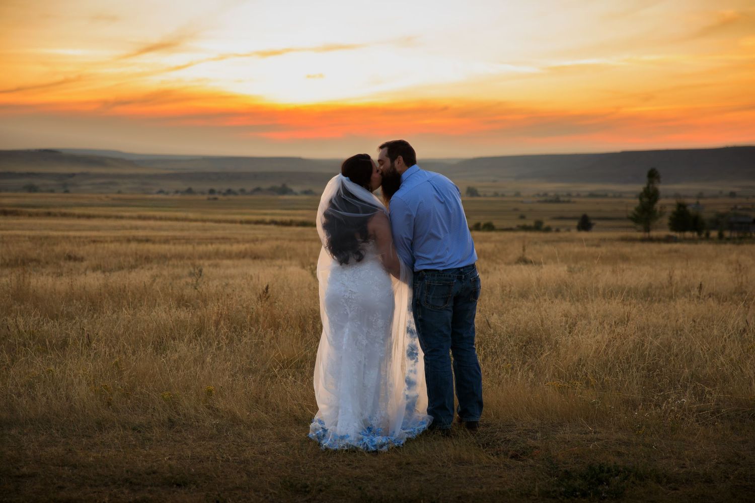 Wedding couple embraces while watching a vibrant orange sunset over golden prairie fields at dusk in Montana.