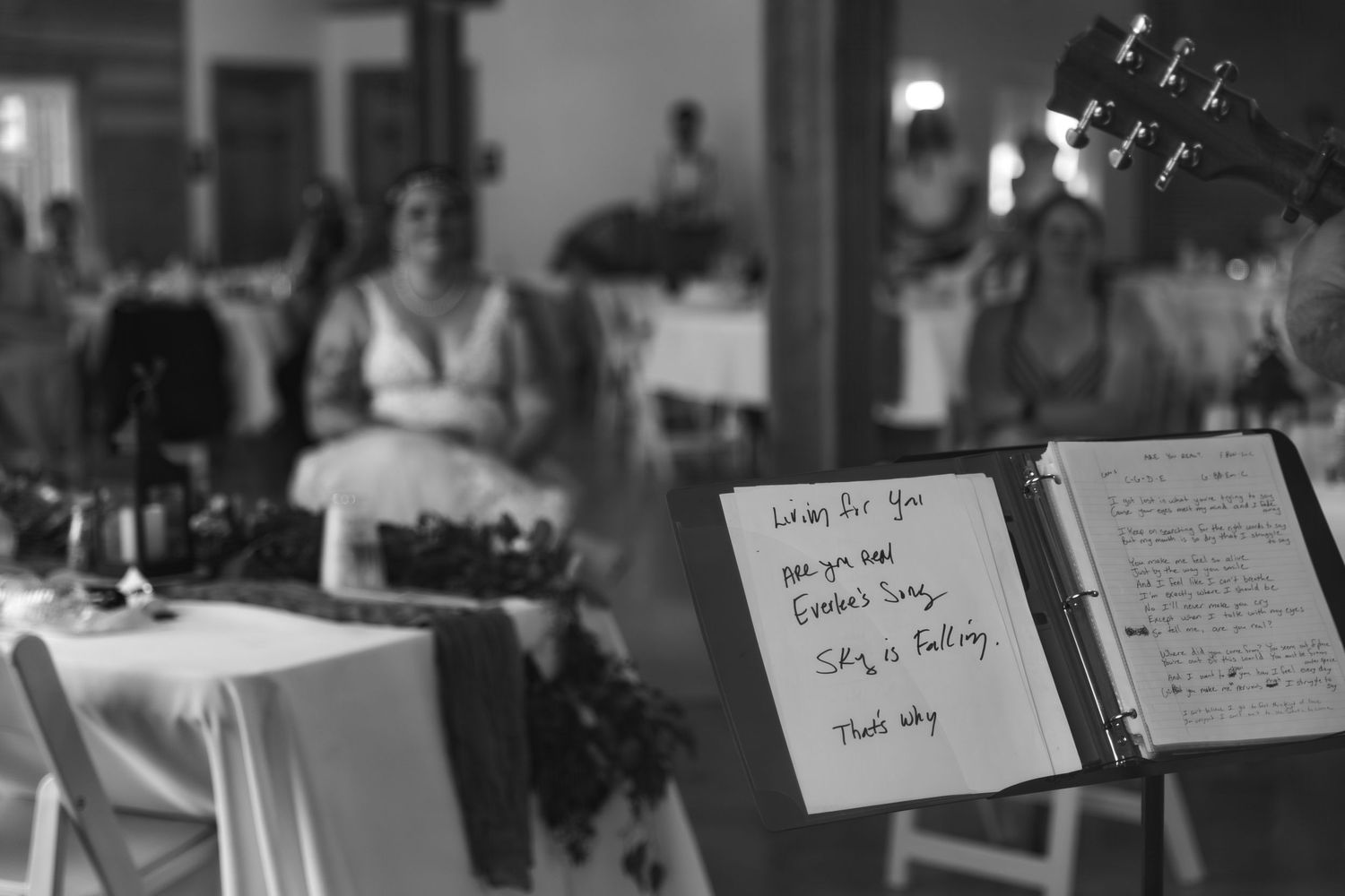 A handwritten menu or note stands in focus while a blurred reception or dining event takes place in the background.
