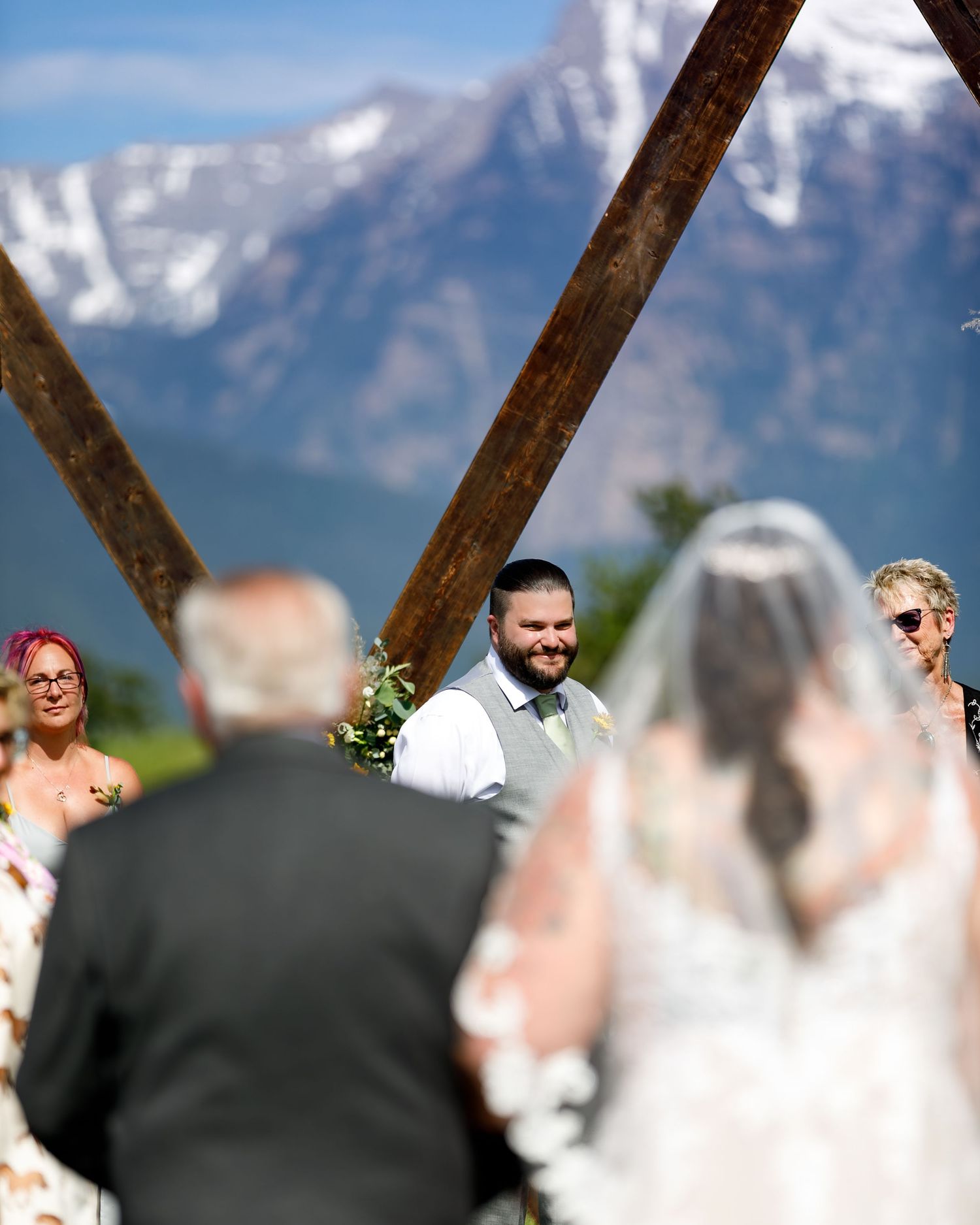 Wedding ceremony framed through wooden beams with snowy mountain peaks in the background.