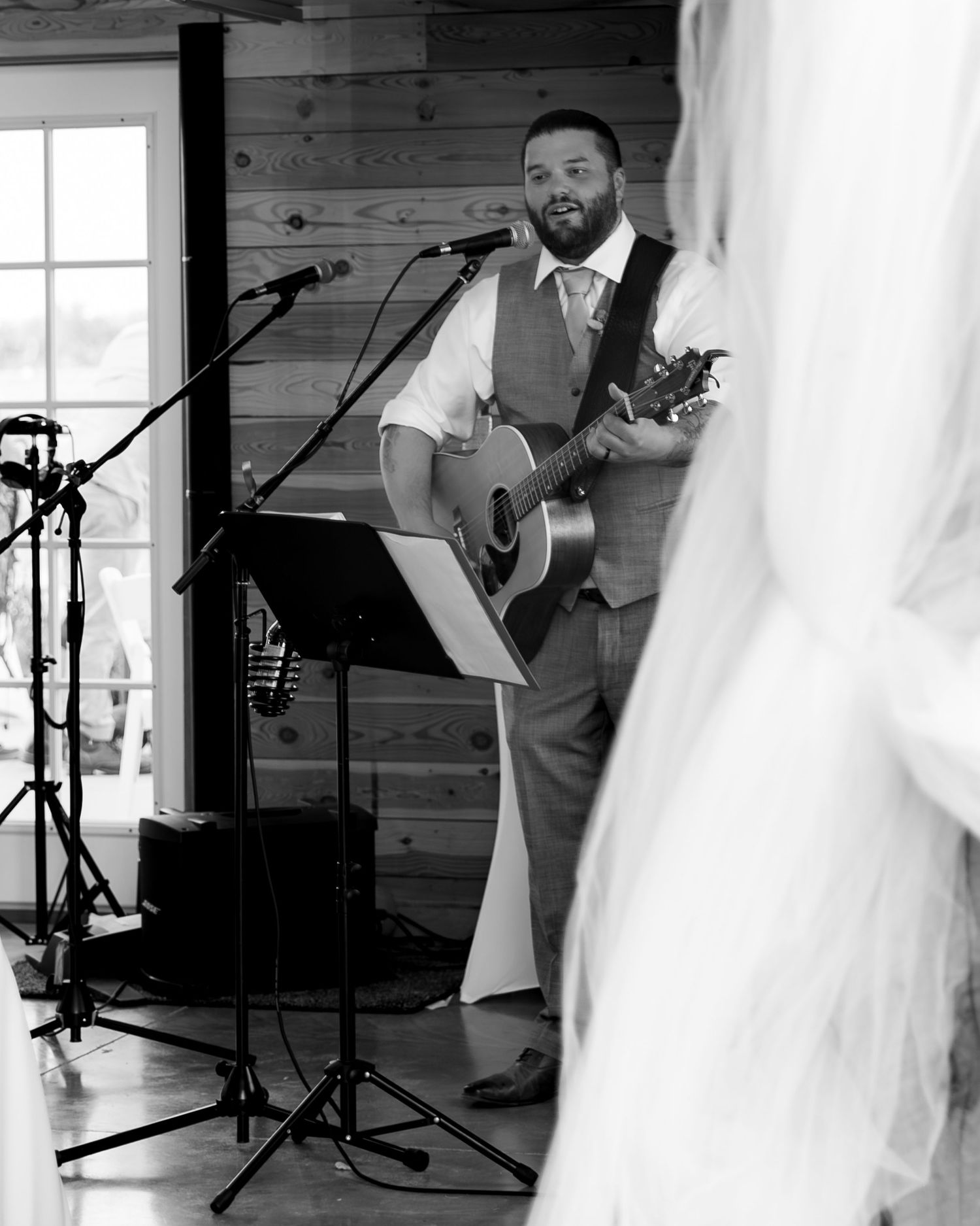 Black and white candid wedding photo showing musicians performing with acoustic guitars in a rustic wooden venue.