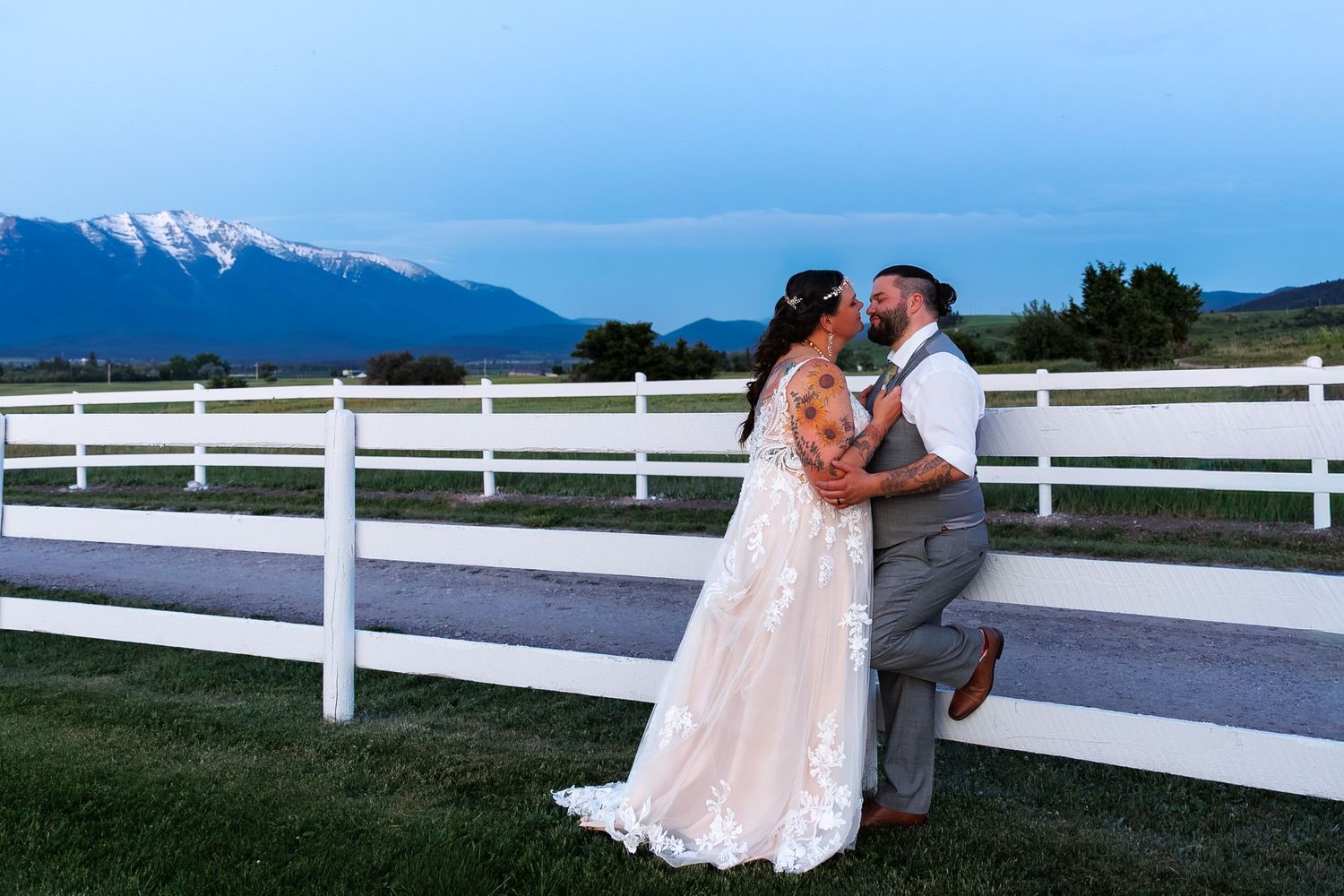 A couple shares a romantic moment by a white fence with mountains in the background at sunset.