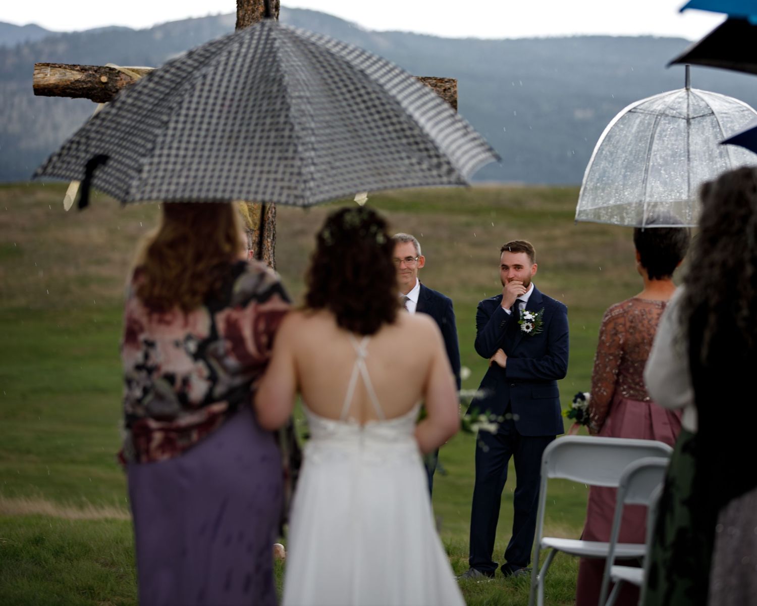 A wedding party stands under umbrellas during a rainy outdoor ceremony in the mountains.
