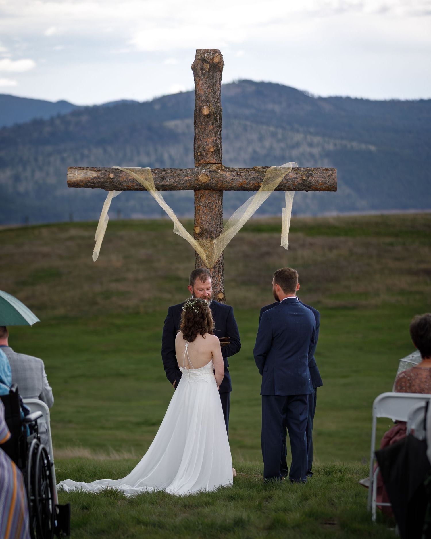 A rustic wooden cross stands against a scenic mountain landscape during an outdoor wedding ceremony.
