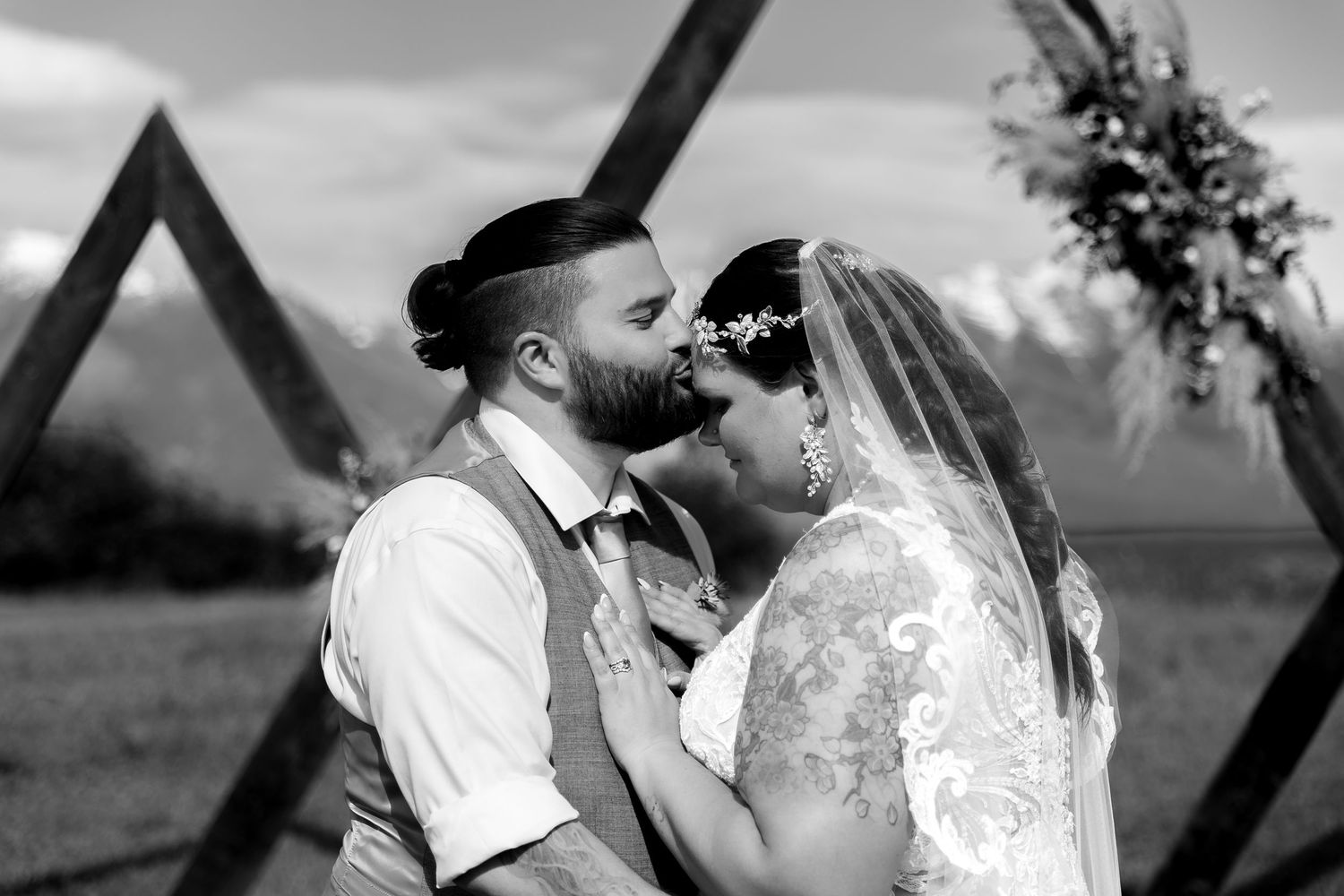 Black and white photo of couple sharing intimate moment under triangular wedding arch during outdoor ceremony.