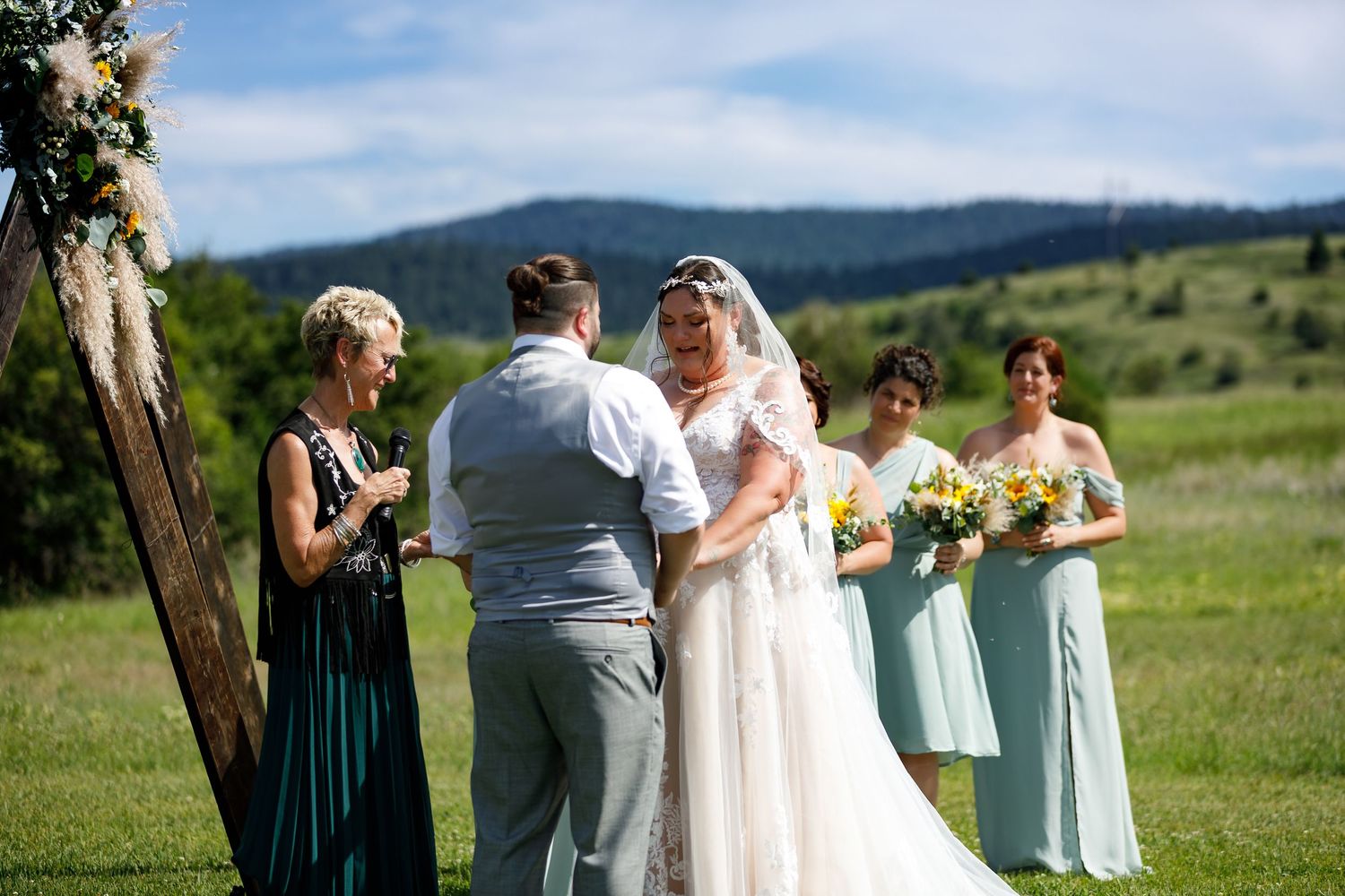 A scenic outdoor wedding ceremony takes place in a green field with mountains in the background.