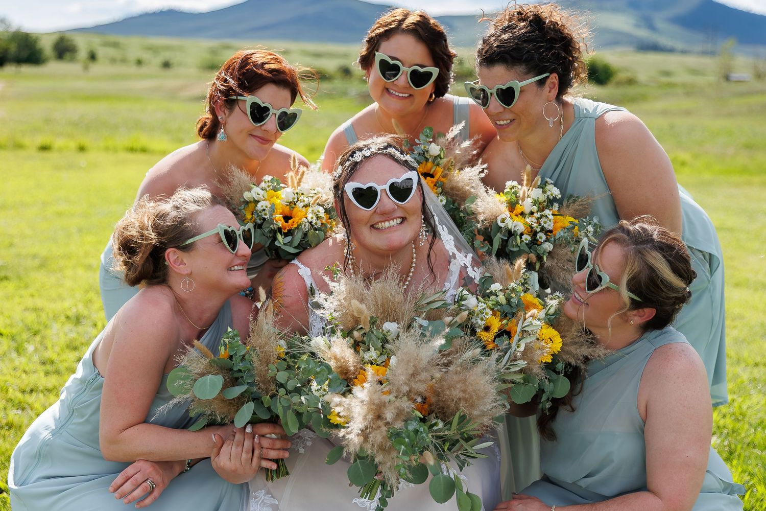 A group of people in mint green dresses and sunglasses pose together with yellow and white floral bouquets in a sunny field.