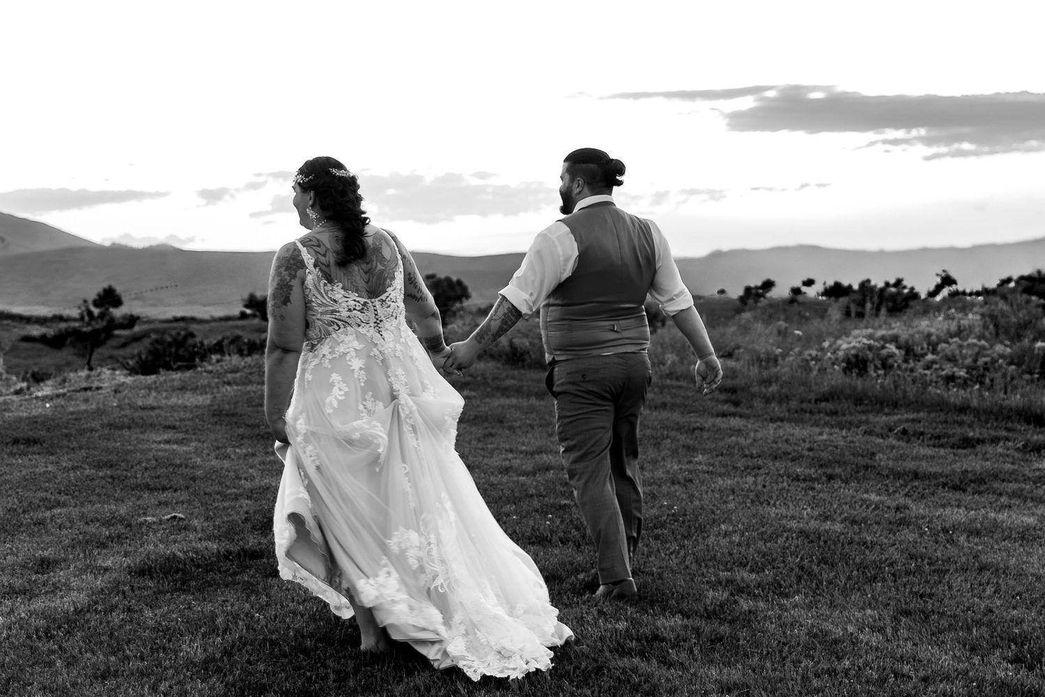 A couple in wedding attire walks hand in hand across a hilltop meadow at sunset in black and white.