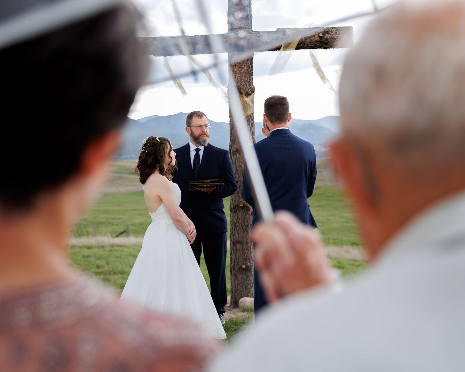 An outdoor wedding ceremony takes place under a wooden arbor with guests watching from their seats.