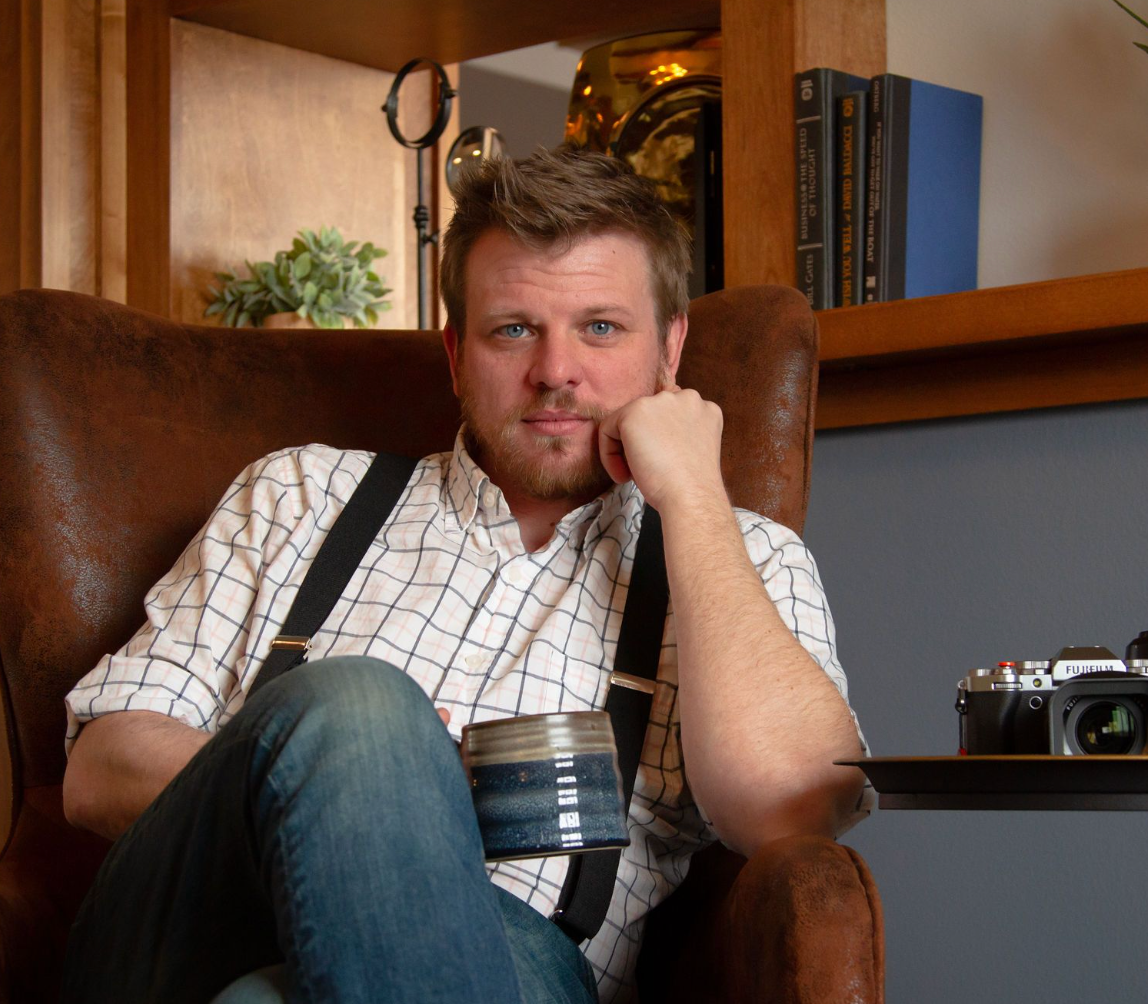 A person in a white checkered shirt sits pensively in a corner against wooden walls and bookcases.