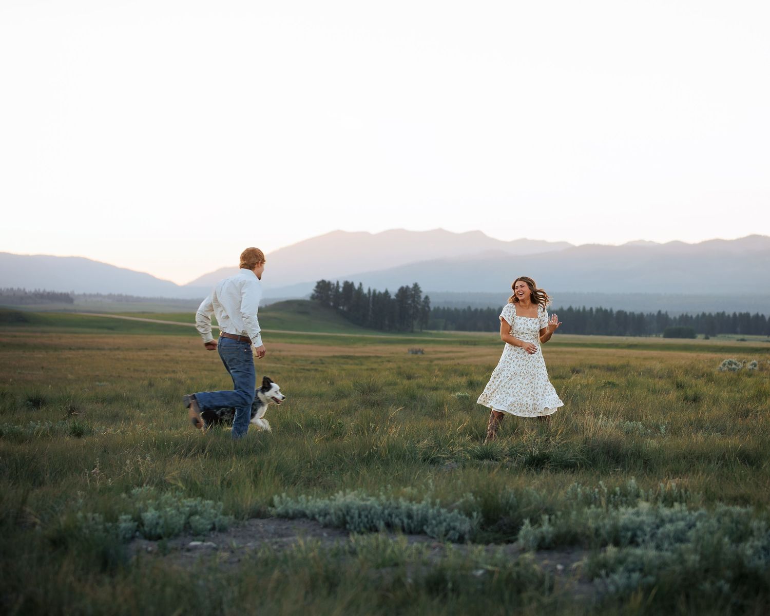 A couple in wedding attire runs playfully through a scenic mountain meadow at dusk.