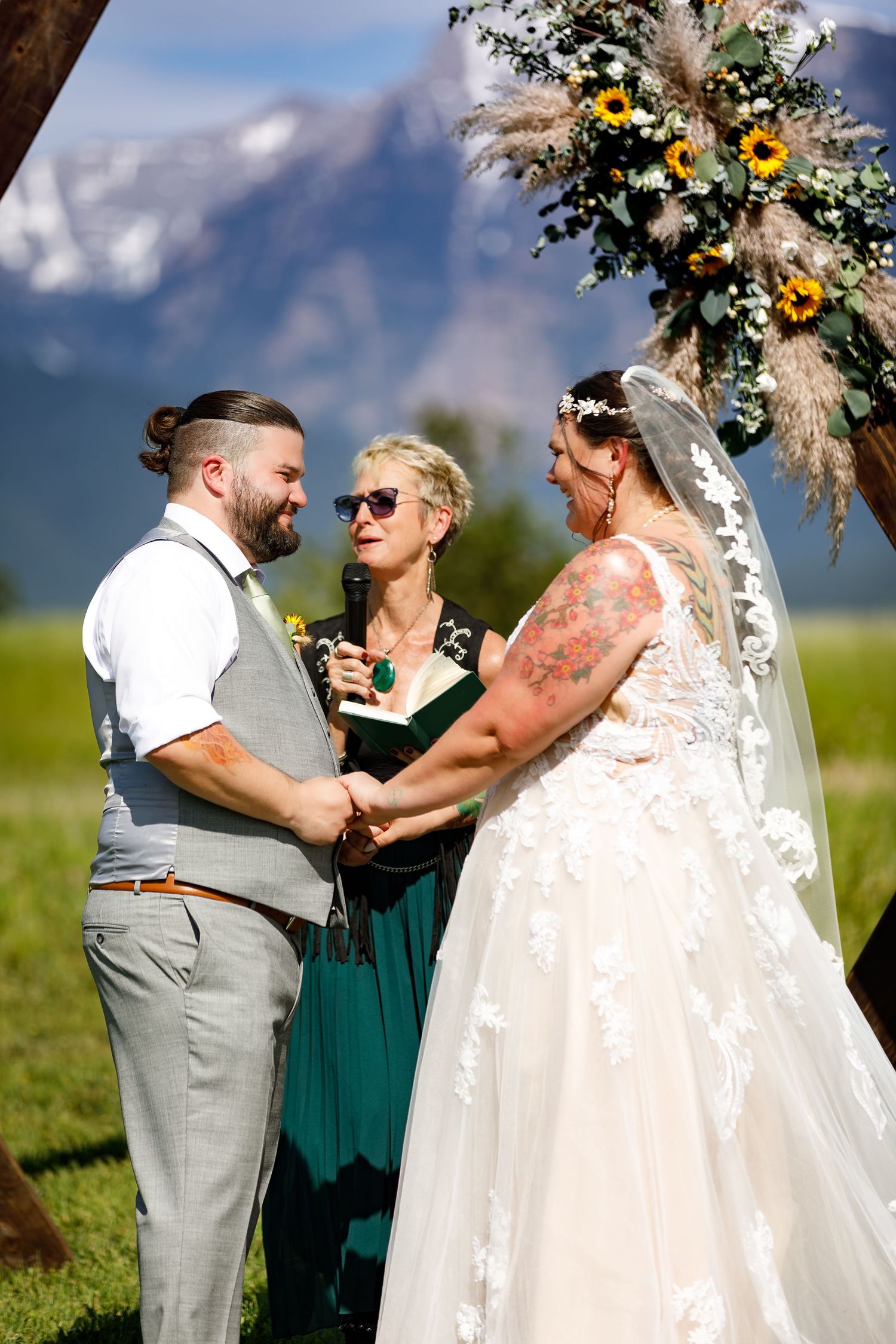 Wedding ceremony officiant in green dress interacts with couple during outdoor mountain ceremony.