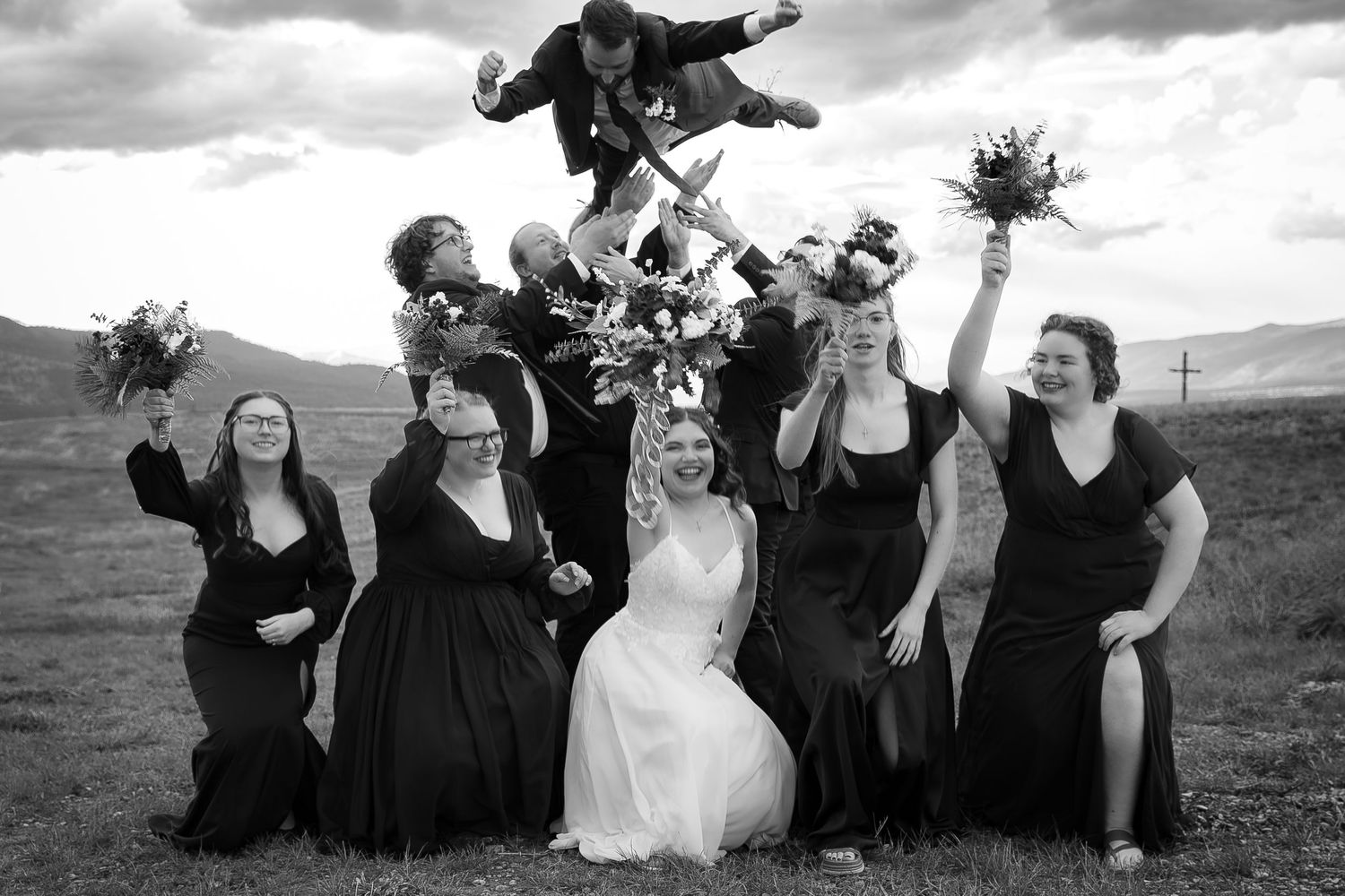A group in black and white formal attire poses playfully outdoors against a cloudy sky backdrop.