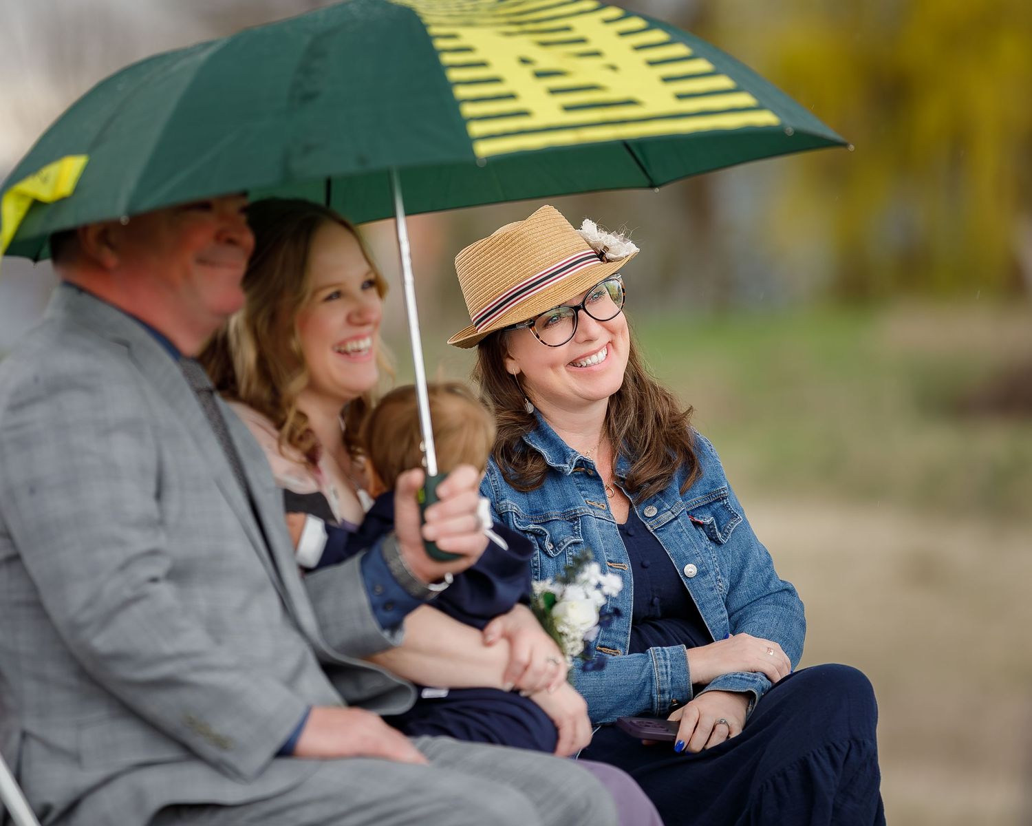 Three people share a green and yellow umbrella while sitting together outdoors on a rainy day.
