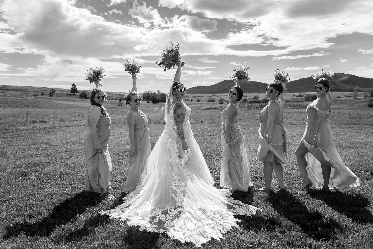 A wedding party in formal attire poses together on a scenic hillside with dramatic clouds overhead.