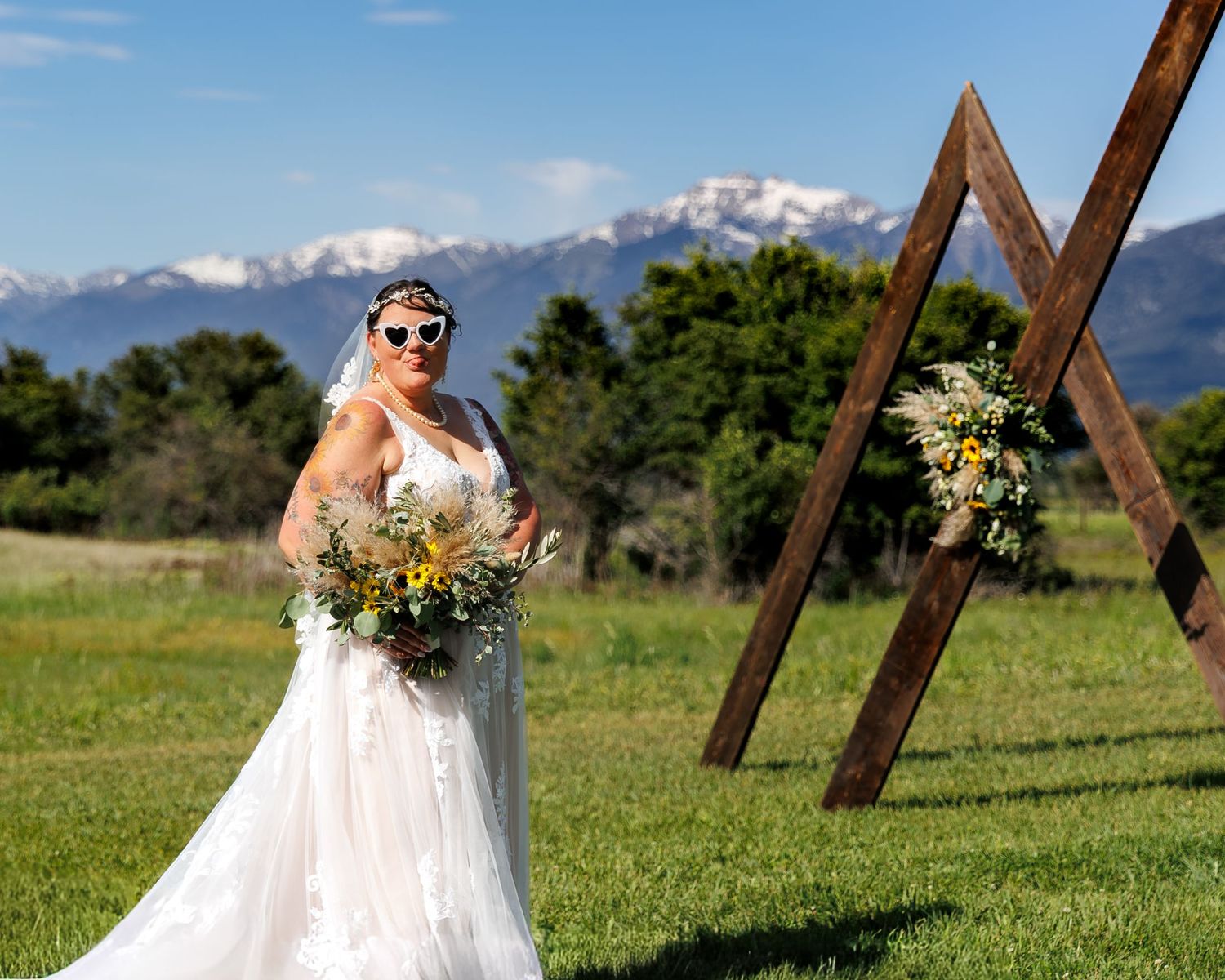 A bride in a white wedding dress holds a colorful bouquet while standing in a grassy field with mountains in the background.