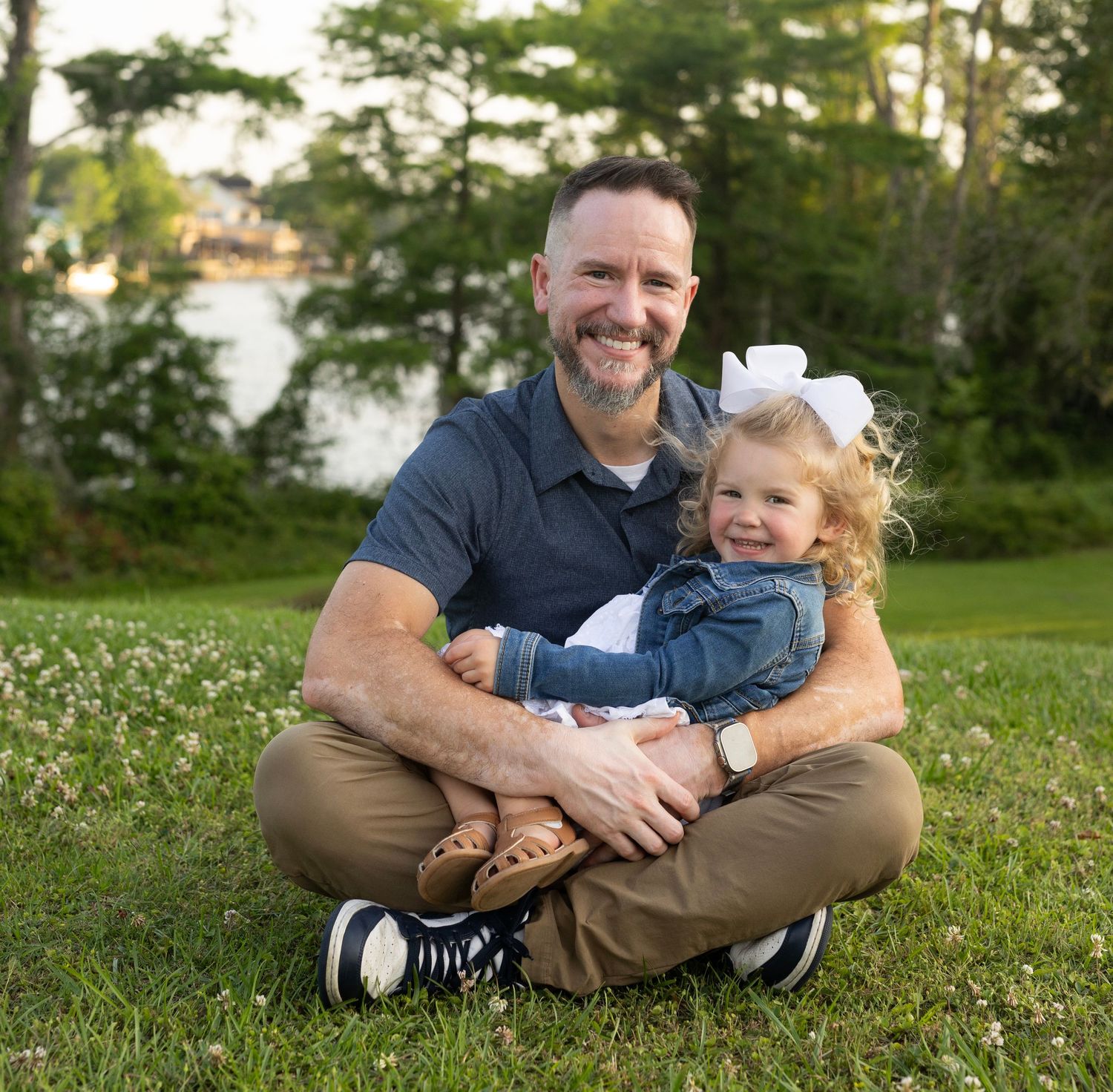 Parent and child sit together on green grass during a sunny outdoor photoshoot near trees and water.