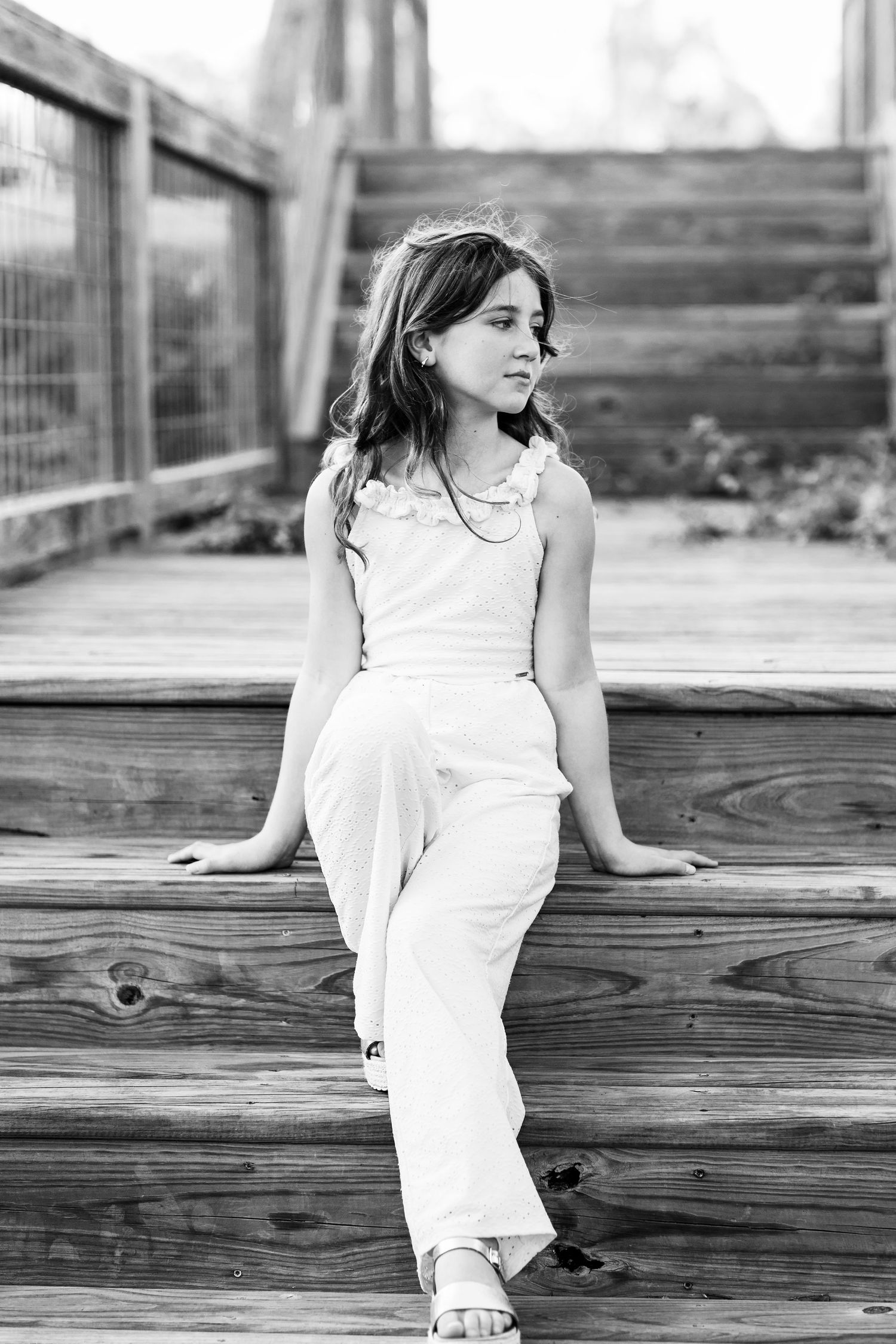 A black and white portrait of a young figure in white clothing sitting pensively on wooden steps outdoors.