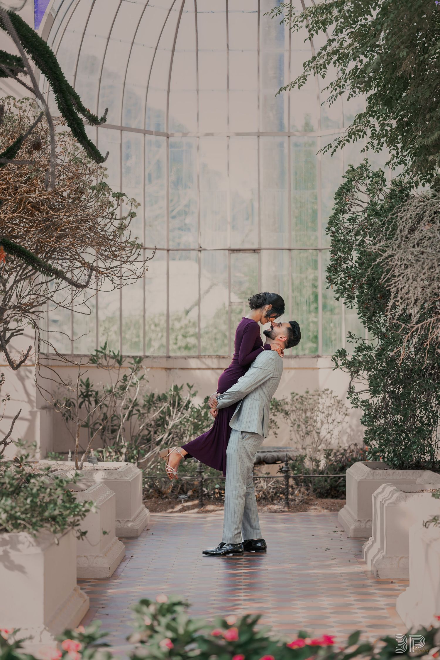 A couple shares a romantic kiss in front of a stone wall with vines and flowers in an outdoor garden setting.
