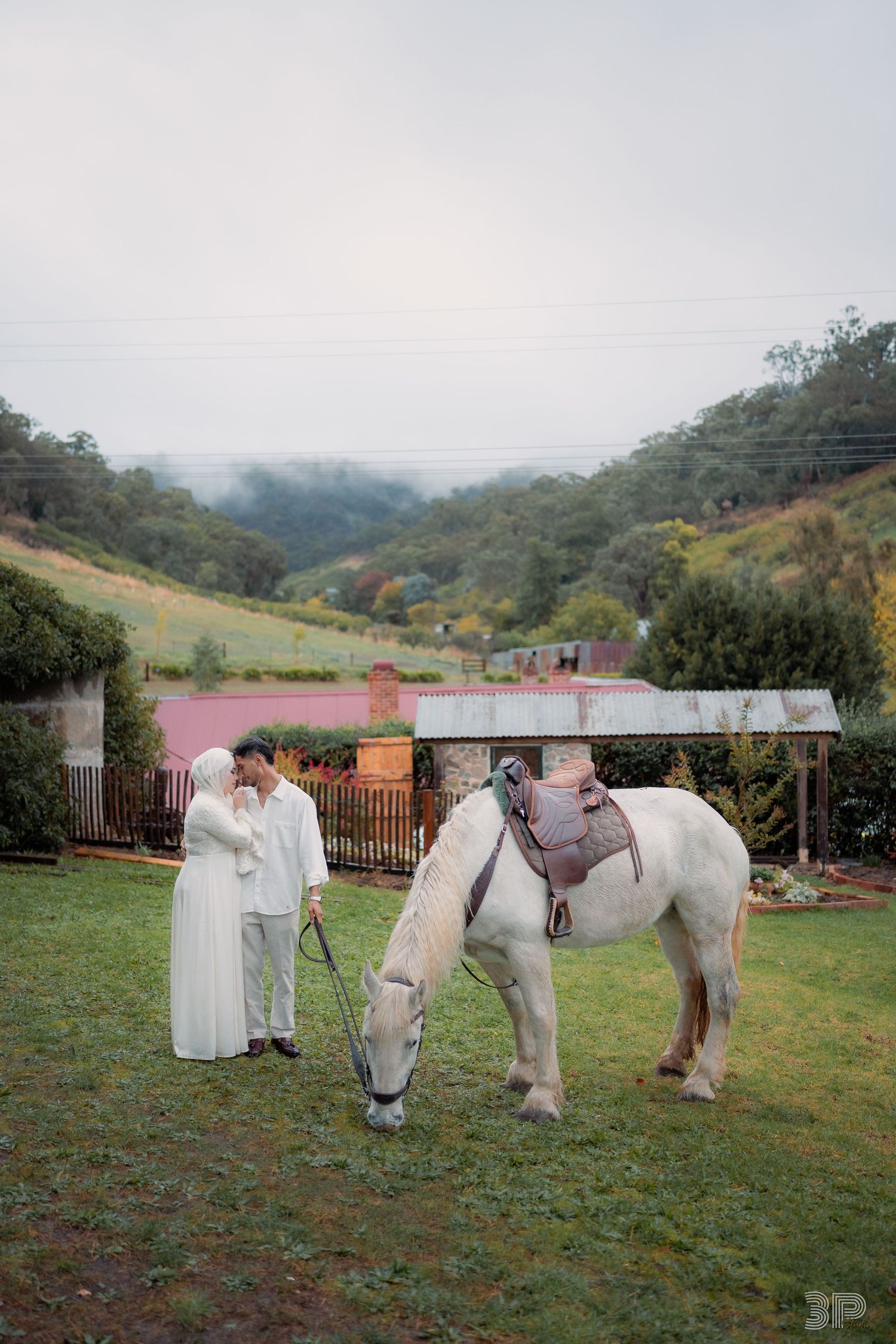 Couple in white attire standing with a white horse on green lawn, rustic buildings and misty hills in background.