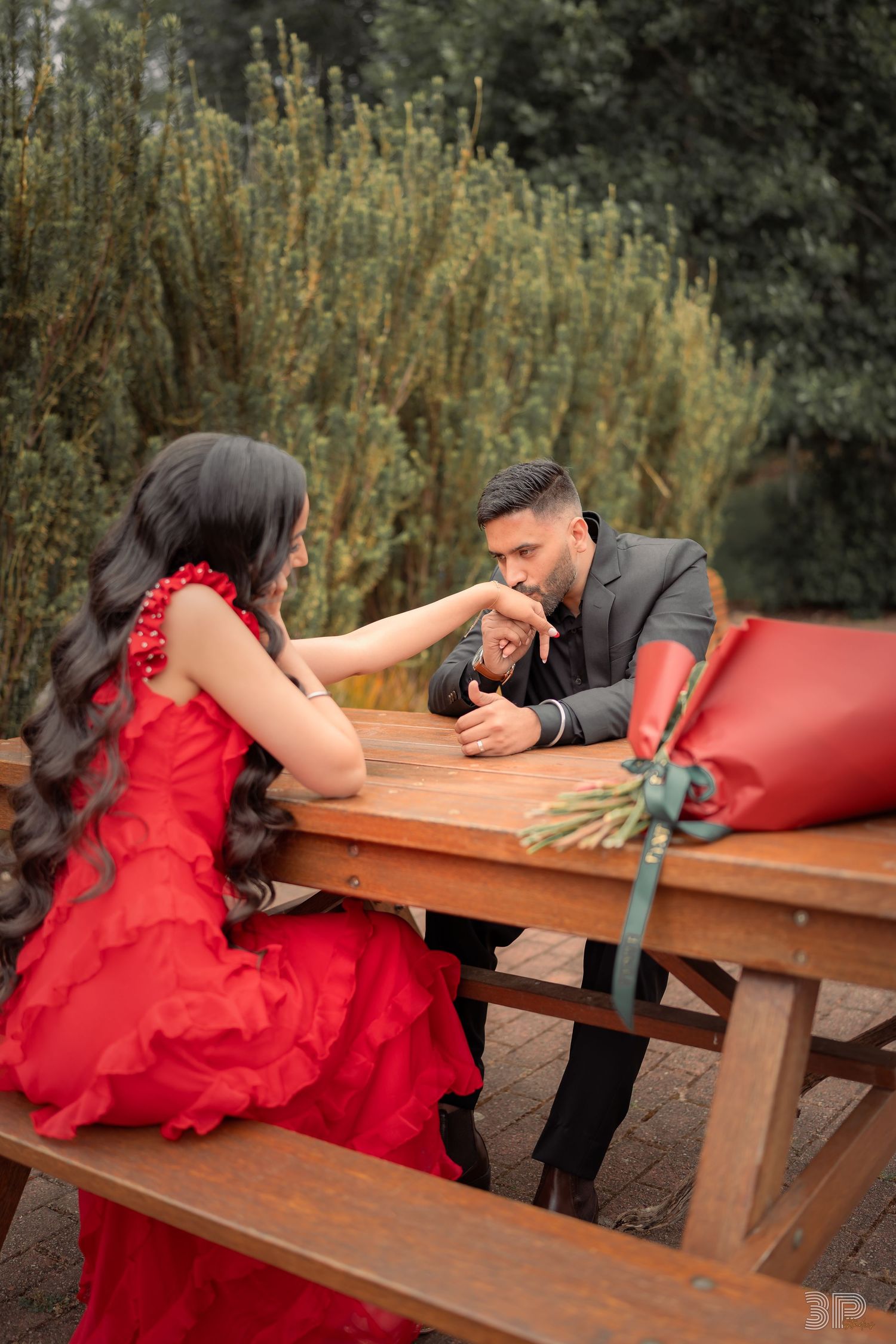 A couple in formal attire playfully interacts across a wooden picnic table in an outdoor setting.