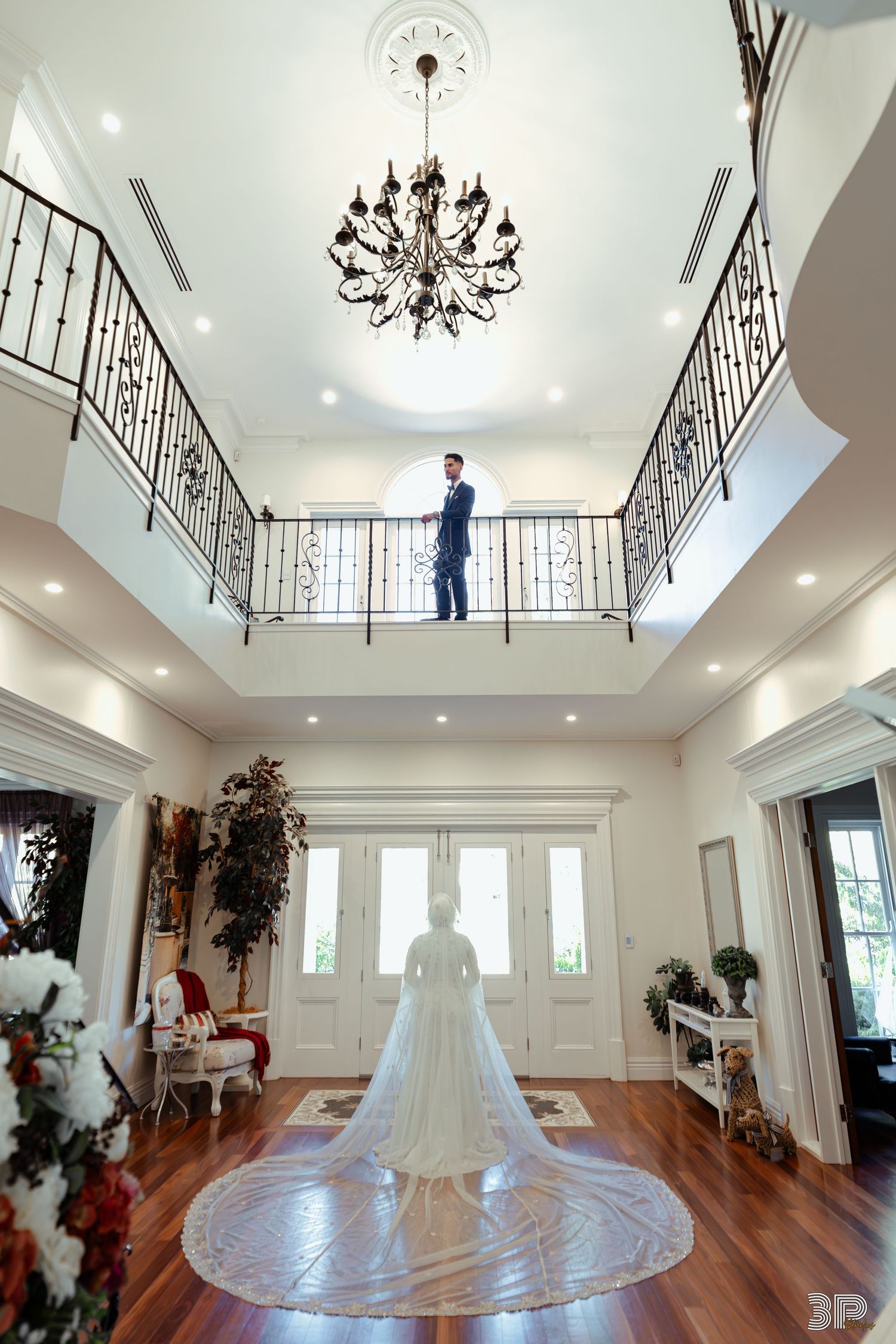 A wedding dress displayed in an elegant white room with hardwood floors and wrought iron railings on the upper level.