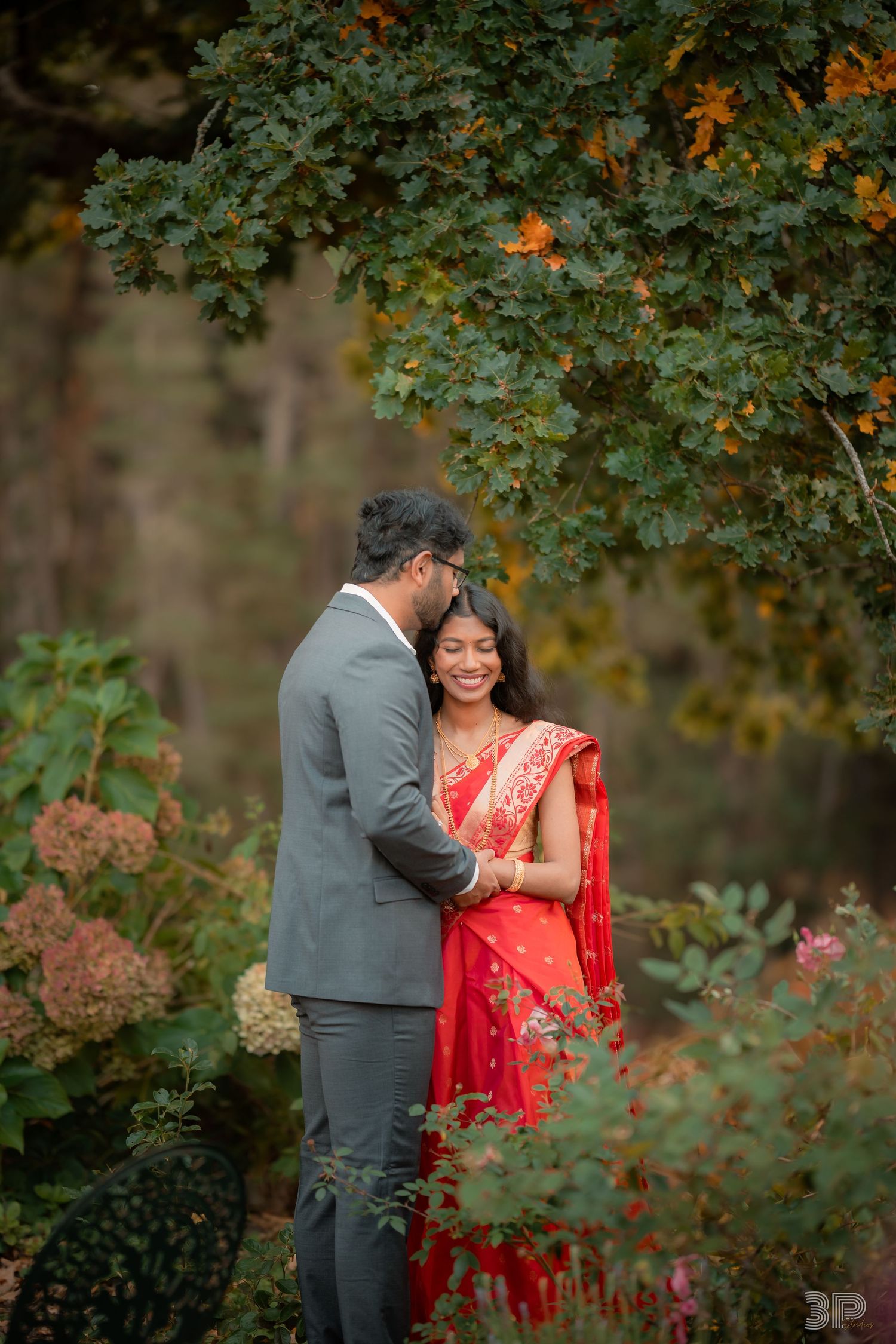 Couple in traditional Indian attire embracing in a lush garden with autumn foliage backdrop.