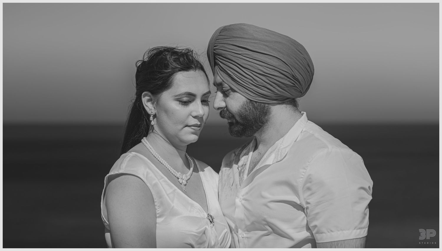 An intimate black and white portrait of a couple in traditional dress against an ocean horizon.