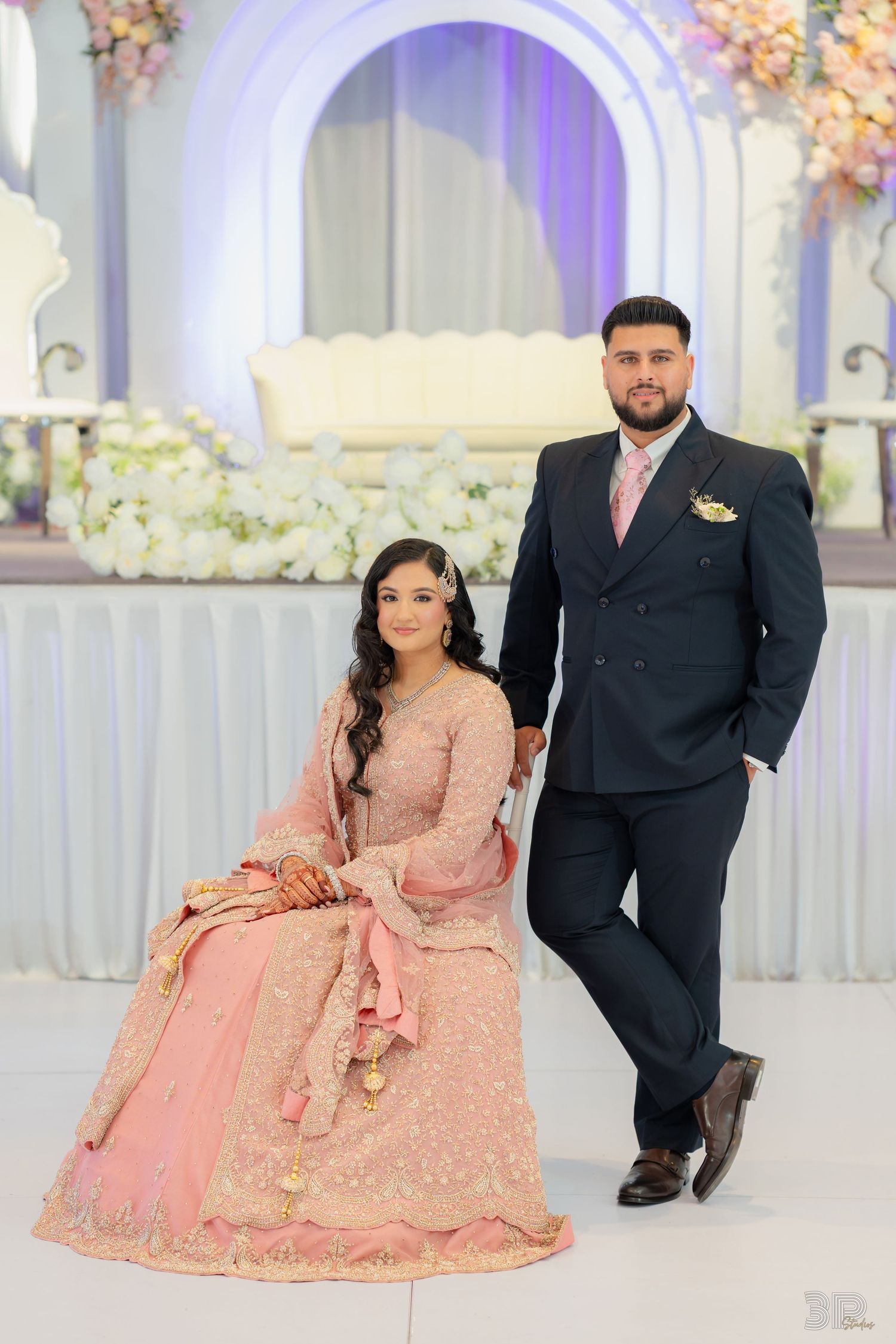 Bride in pink lehenga and groom in navy suit pose elegantly at a decorated wedding venue with floral backdrop.