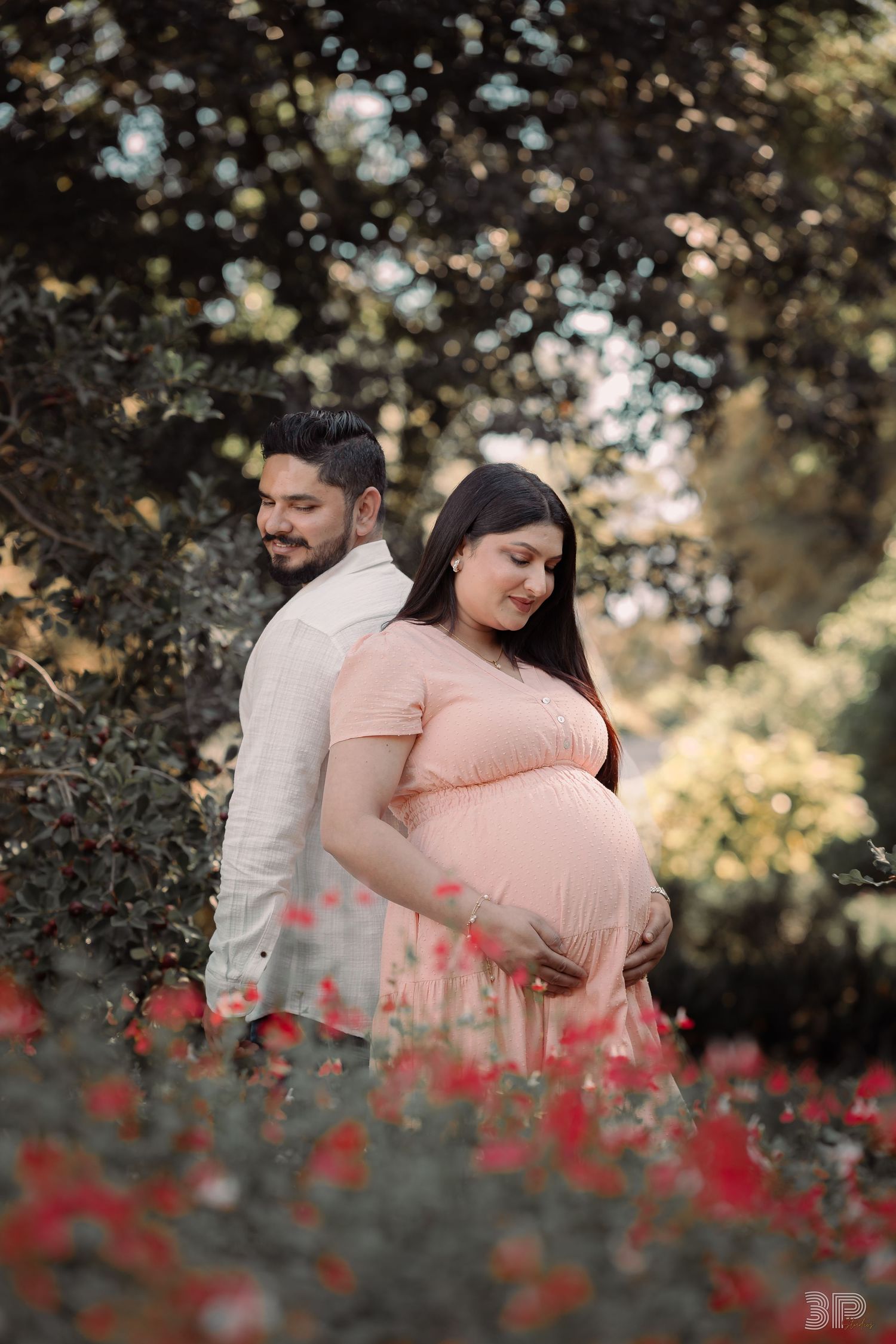 Pregnant woman in pink dress cradling baby bump with partner in garden surrounded by red flowers.