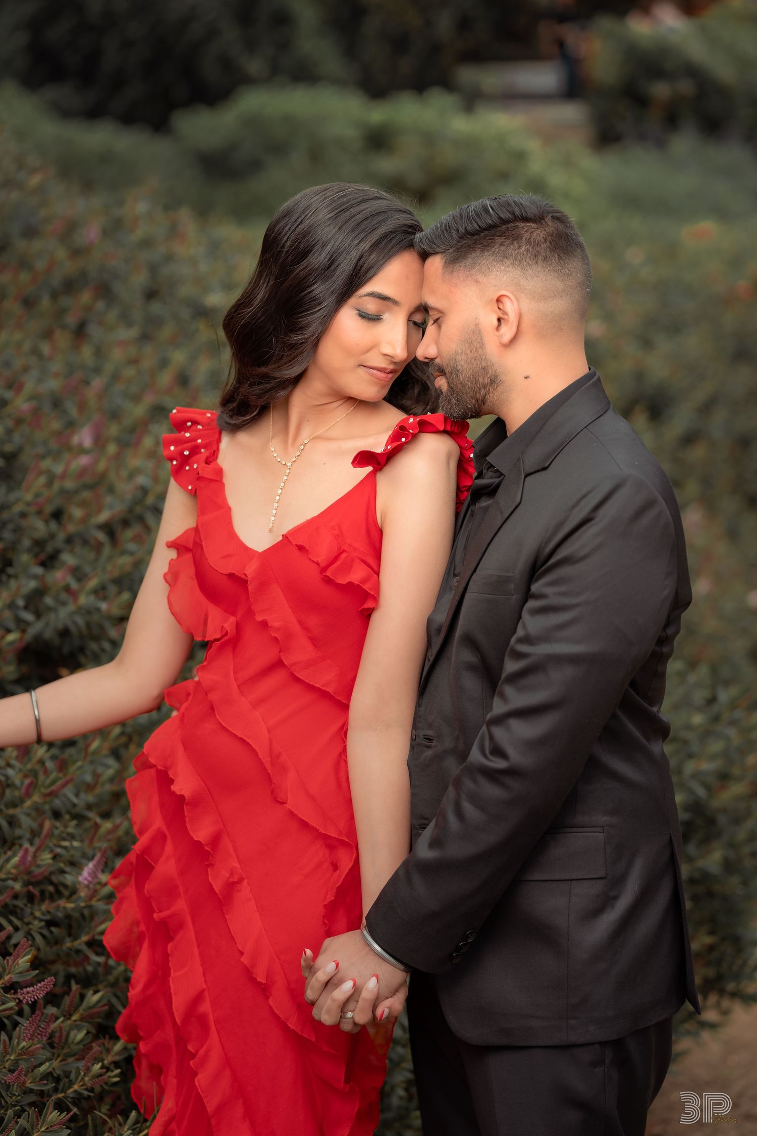 A romantic couple embraces outdoors, with one wearing a flowing red ruffled dress and the other in a dark suit.