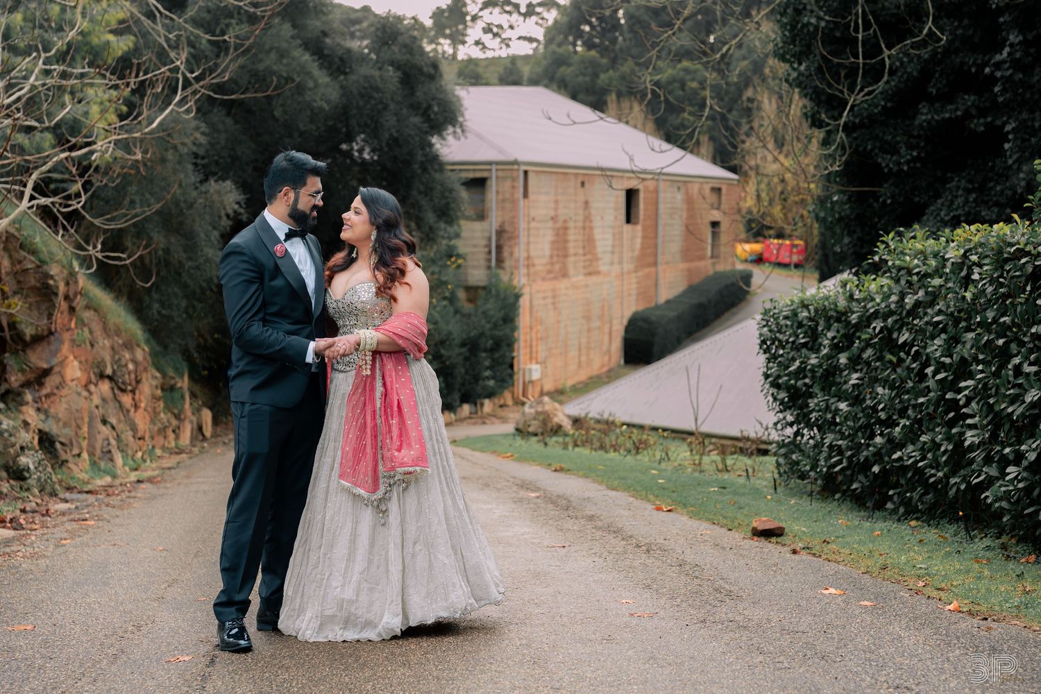 Couple in Indian wedding attire poses on a tree-lined path, man in navy suit, woman in pink and grey lehenga.
