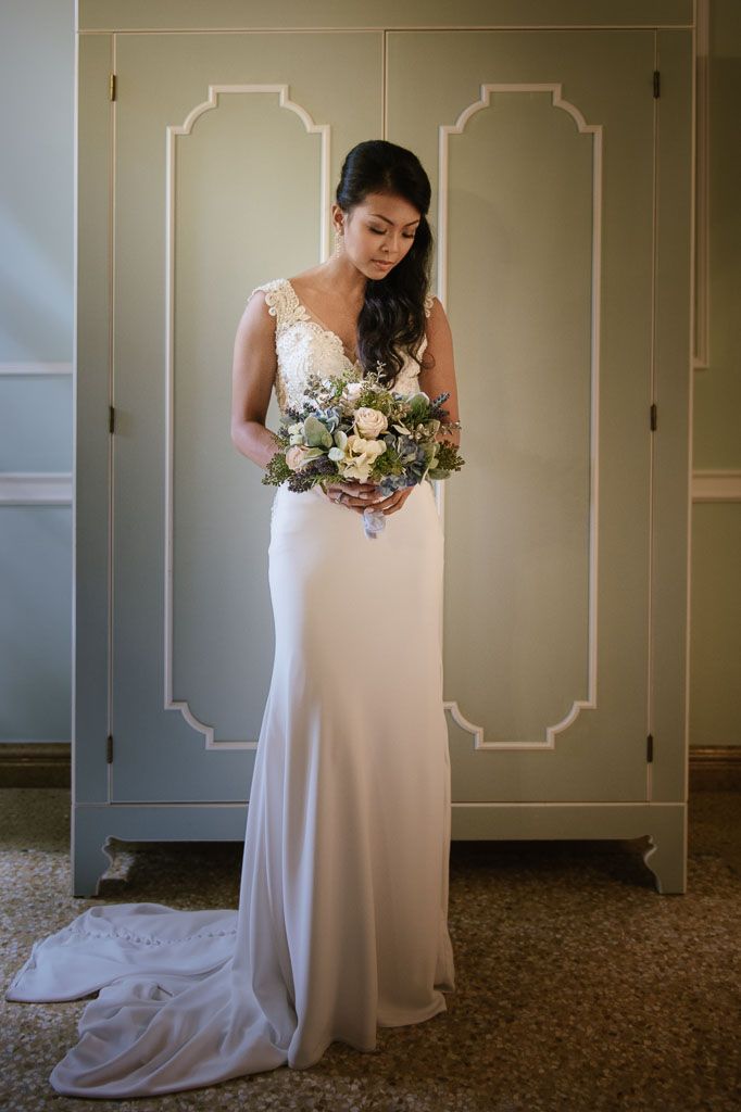 Bride in an elegant white V-neck wedding dress admires her bouquet of white and cream flowers in a hotel hallway.