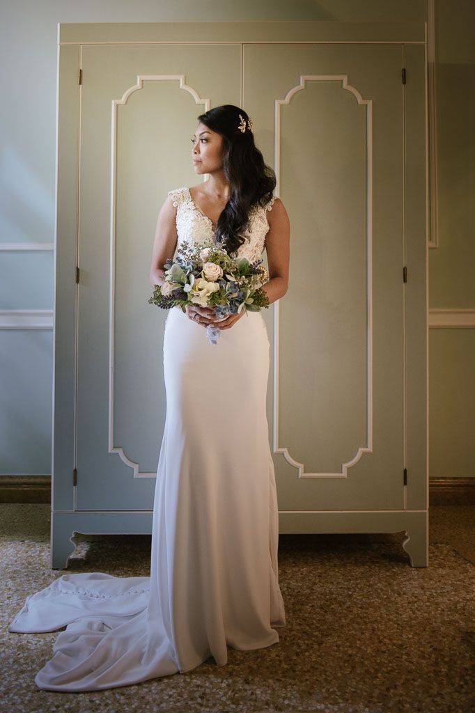A person in an elegant white wedding gown stands gracefully by a decorated wall in an indoor setting.