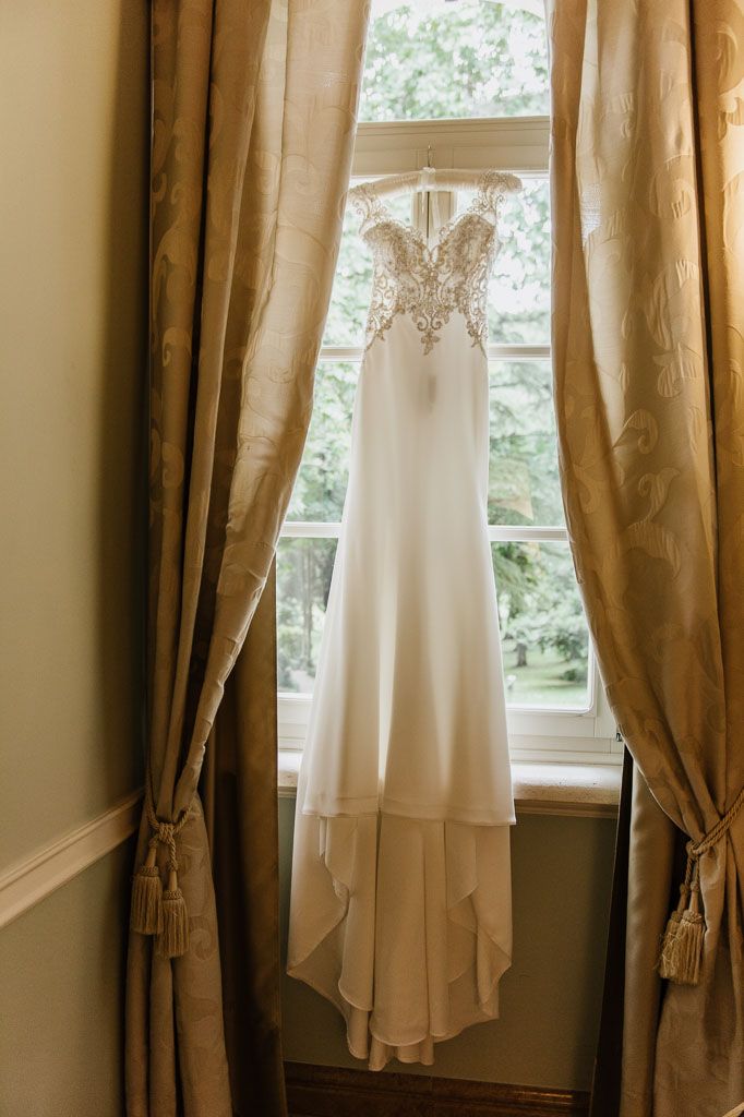 White wedding dress hangs from curtain rod in front of beige drapes and sunlit window.