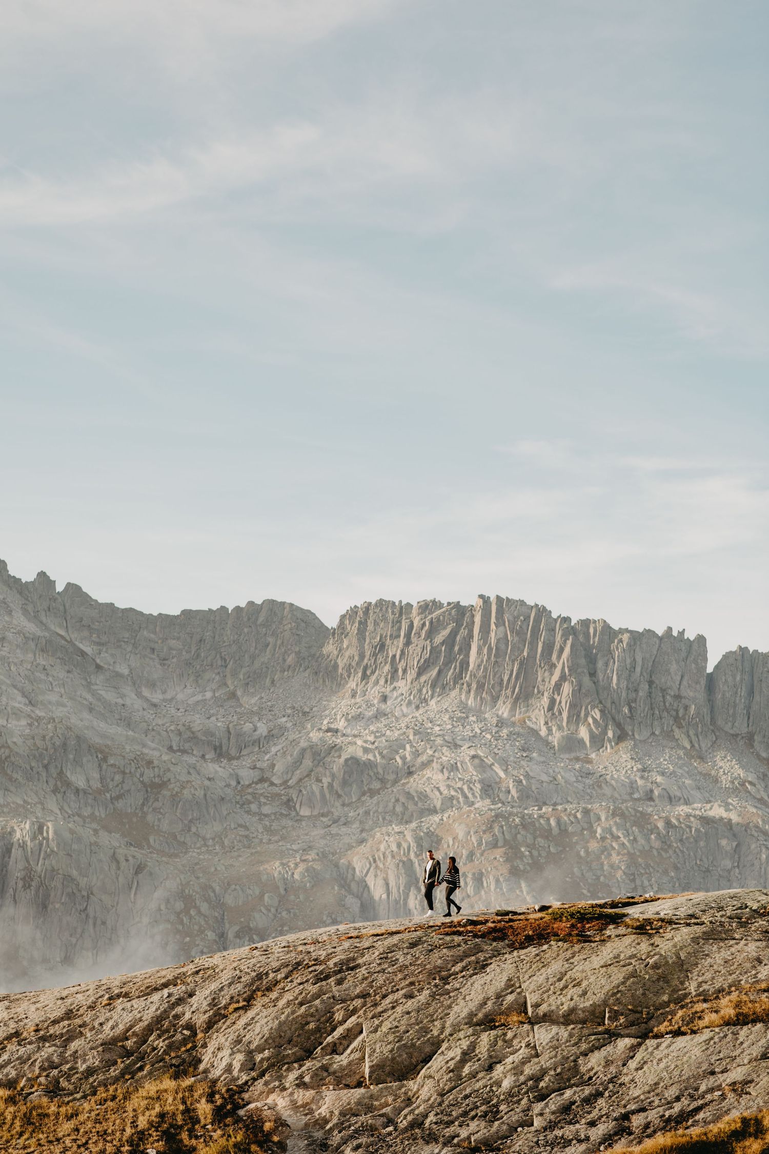 Two tiny silhouettes stand on rocky outcrop beneath dramatic mountain peaks.