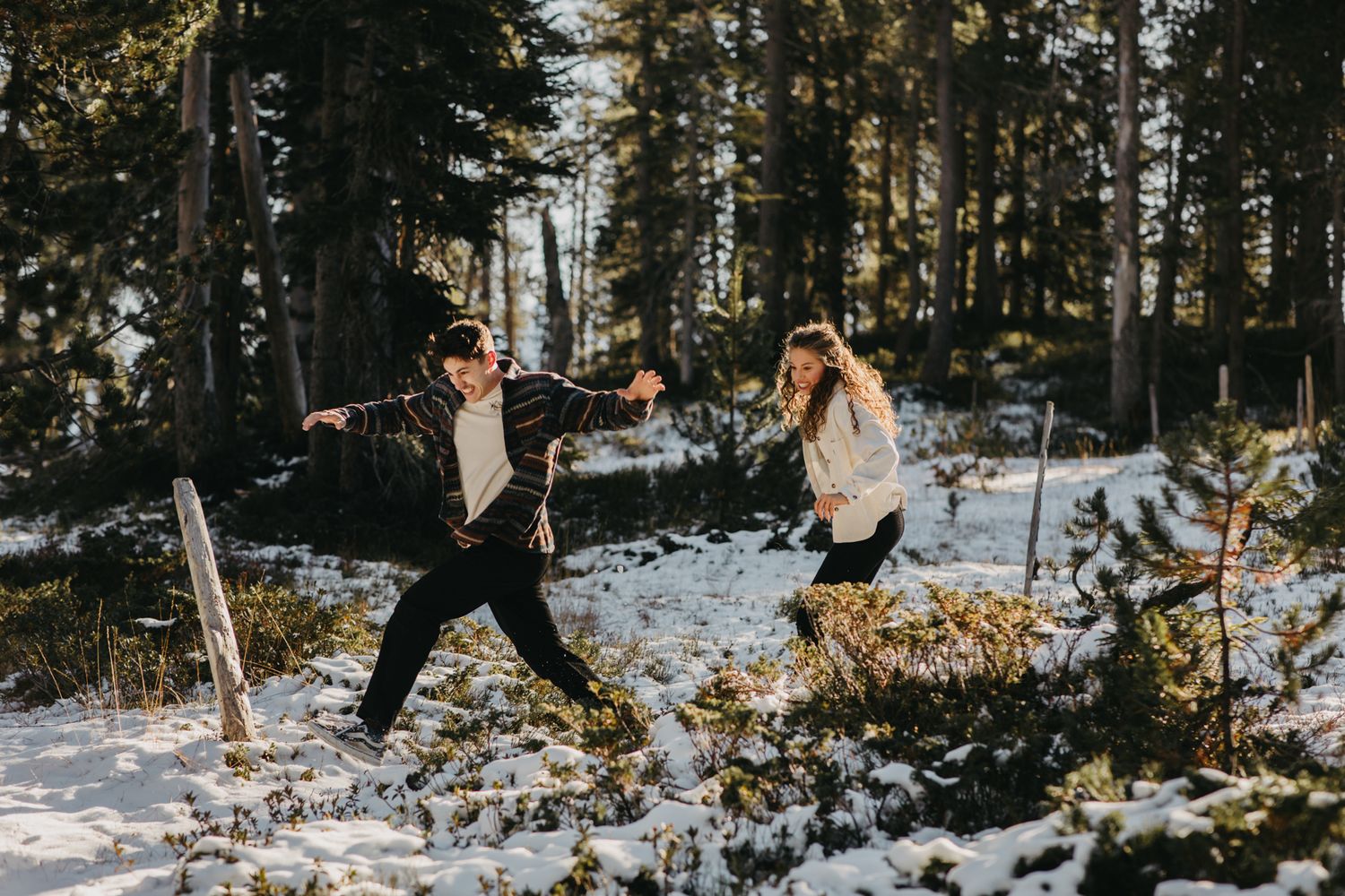 Two people jumping playfully in snow-covered forest during winter photoshoot.