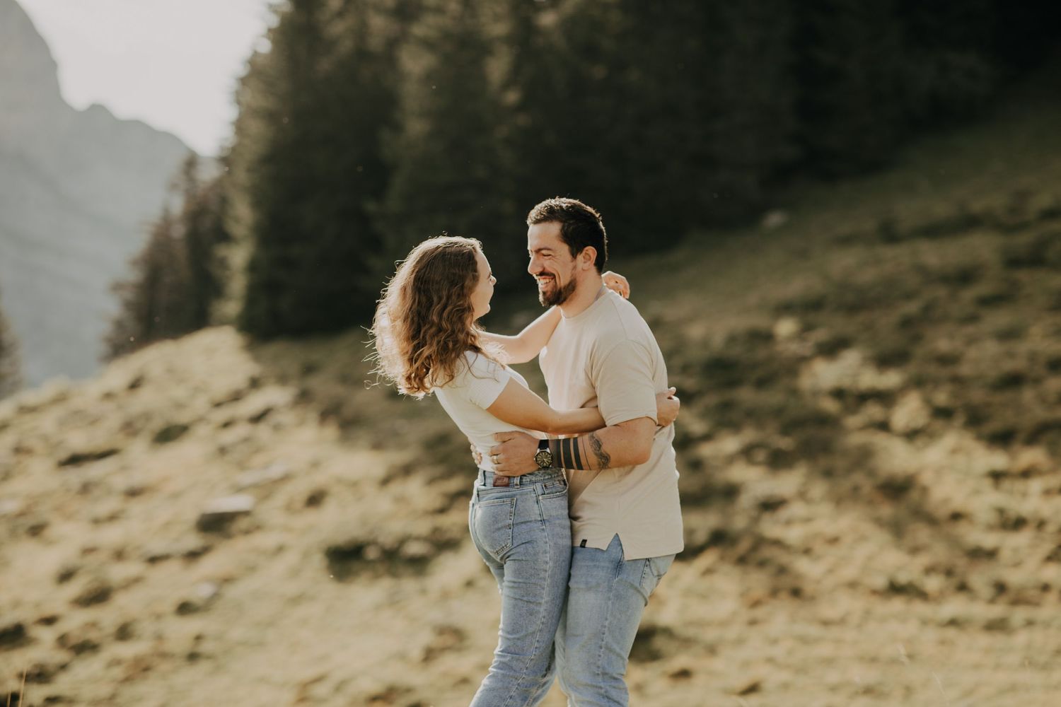 A couple embraces on a sunny mountainside with evergreen trees and golden light in the background.