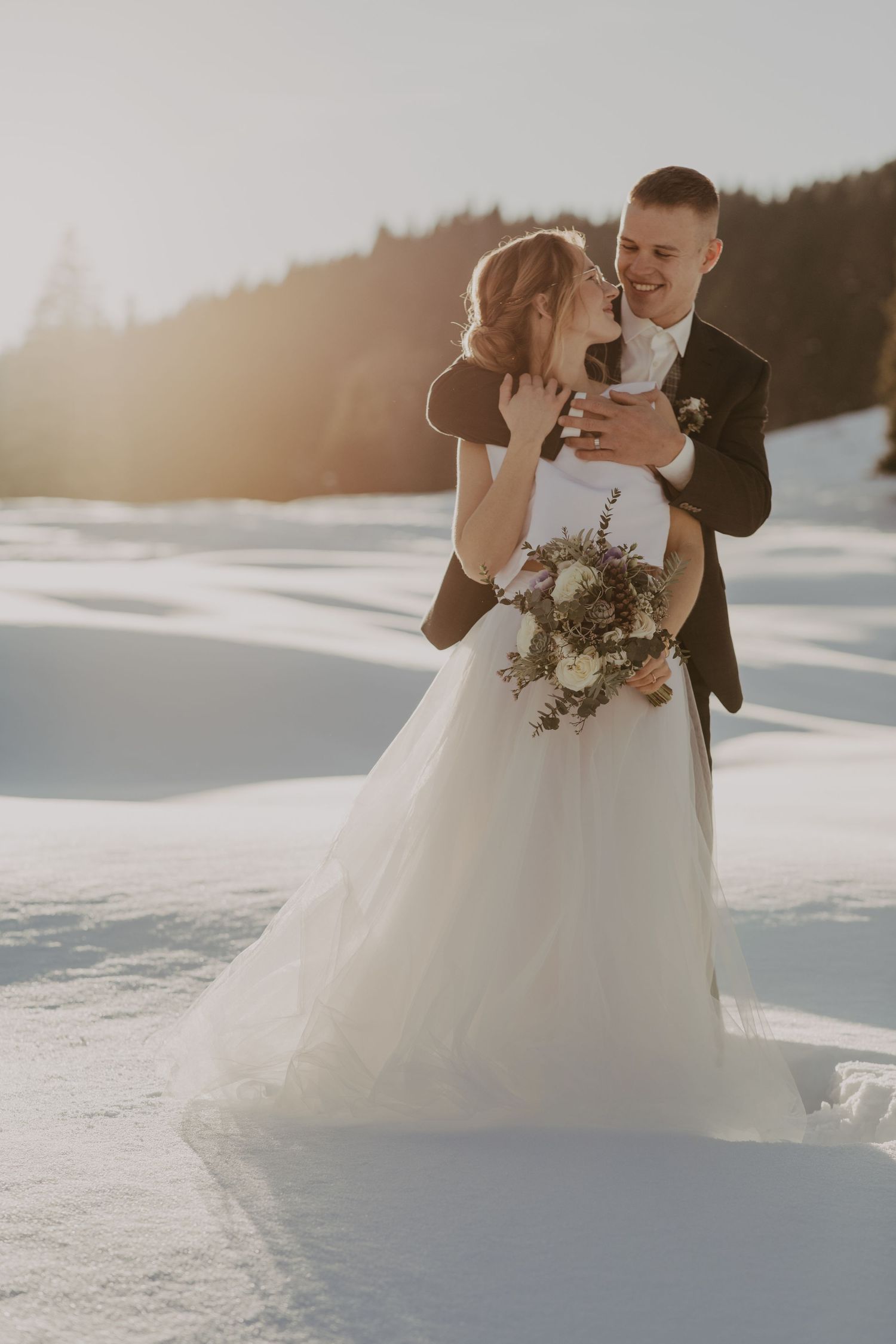Winter wedding couple embracing in snowy mountain landscape at sunset.