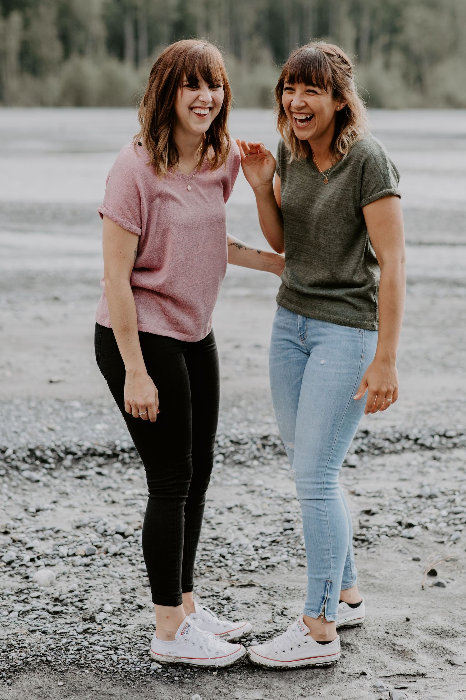 Two friends laughing together on a beach wearing casual outfits and white sneakers.