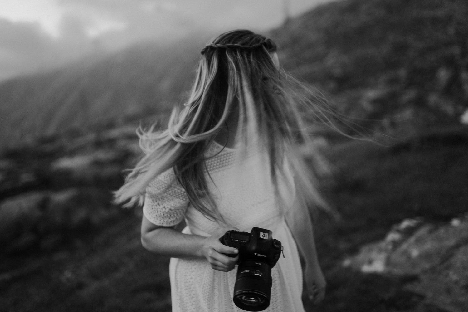 Black and white portrait of photographer with windswept hair holding camera in mountains.