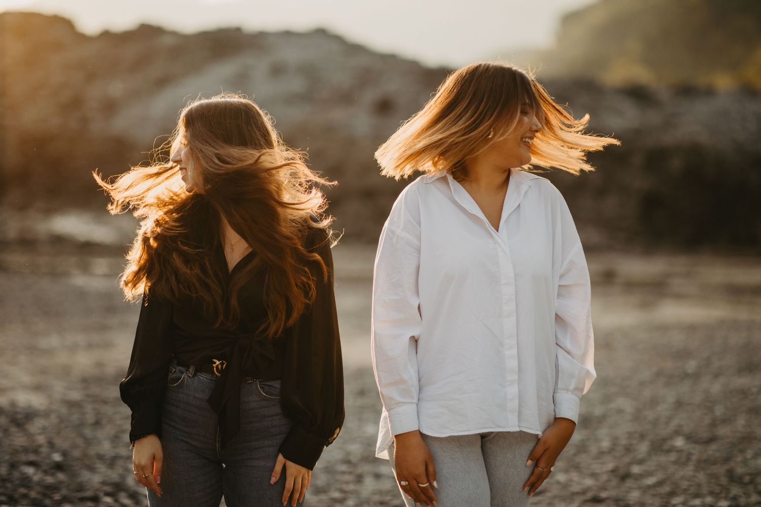 Two women with flowing hair walk along a rocky beach at sunset.