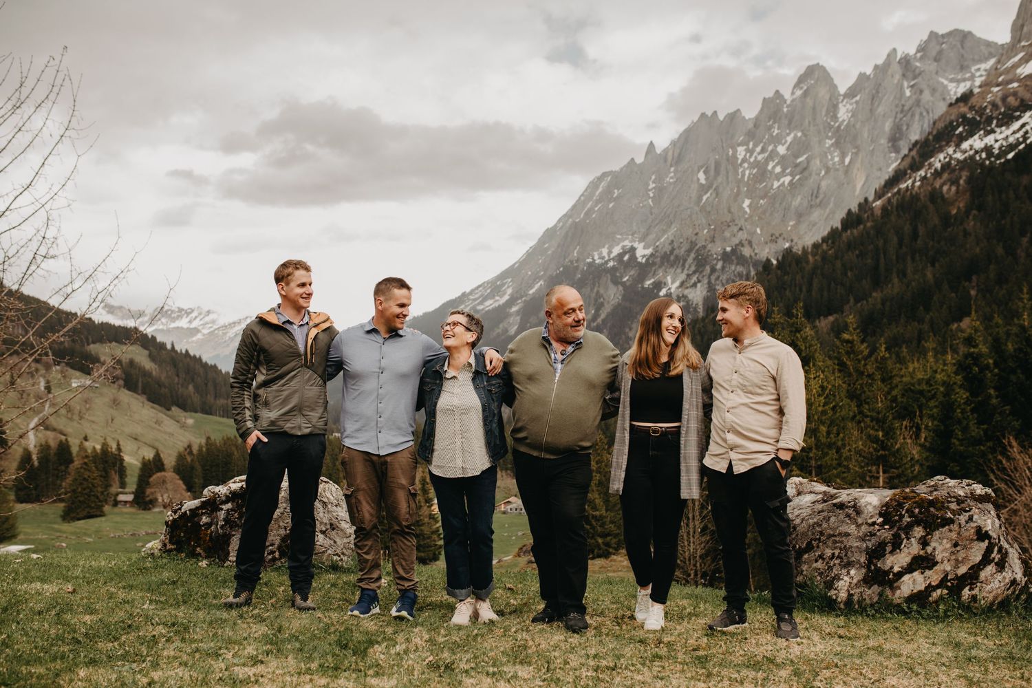 A group of people stand together smiling against a backdrop of majestic alpine mountains in a scenic meadow.