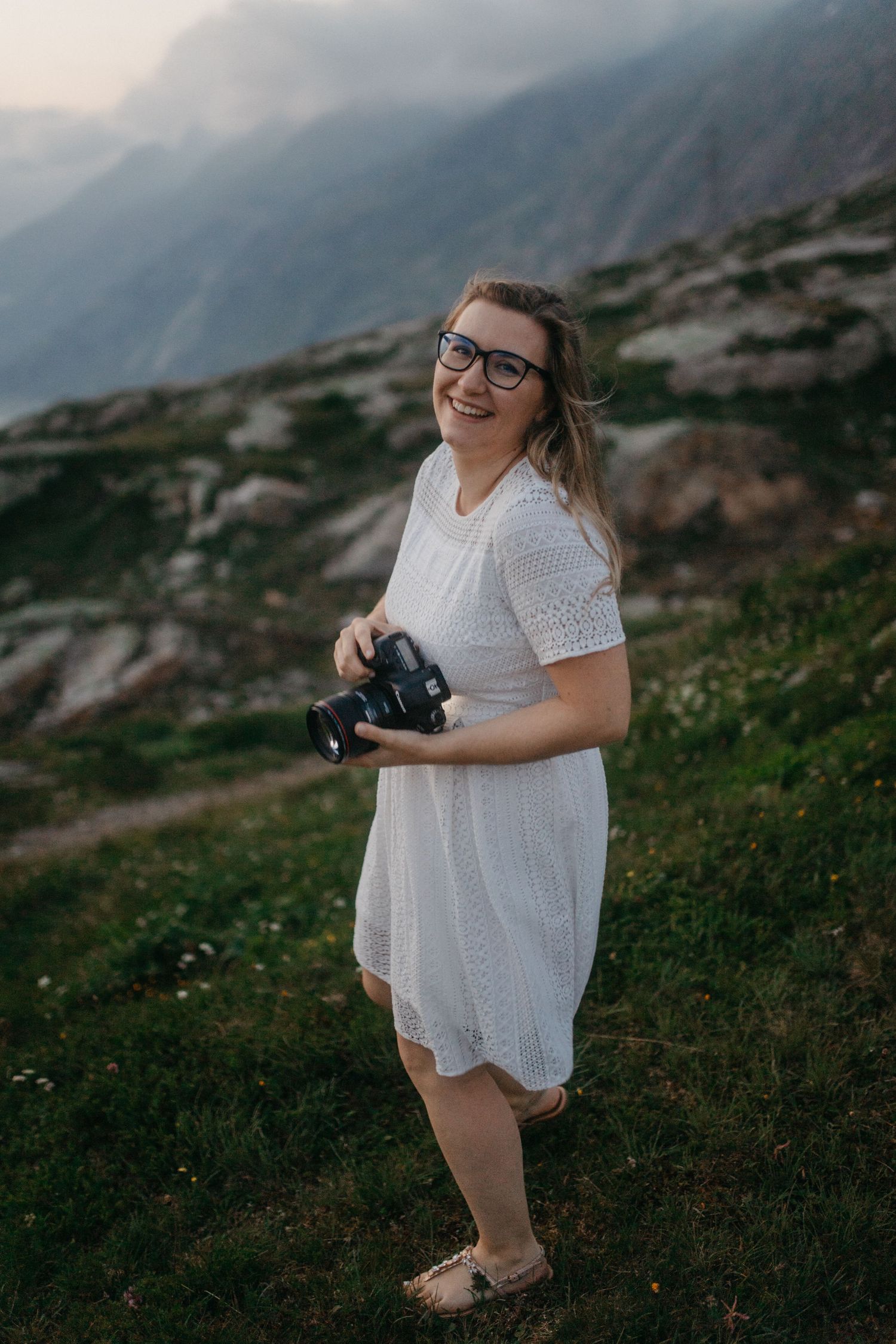 Photographer in white lace dress and glasses poses with camera against mountainous backdrop.